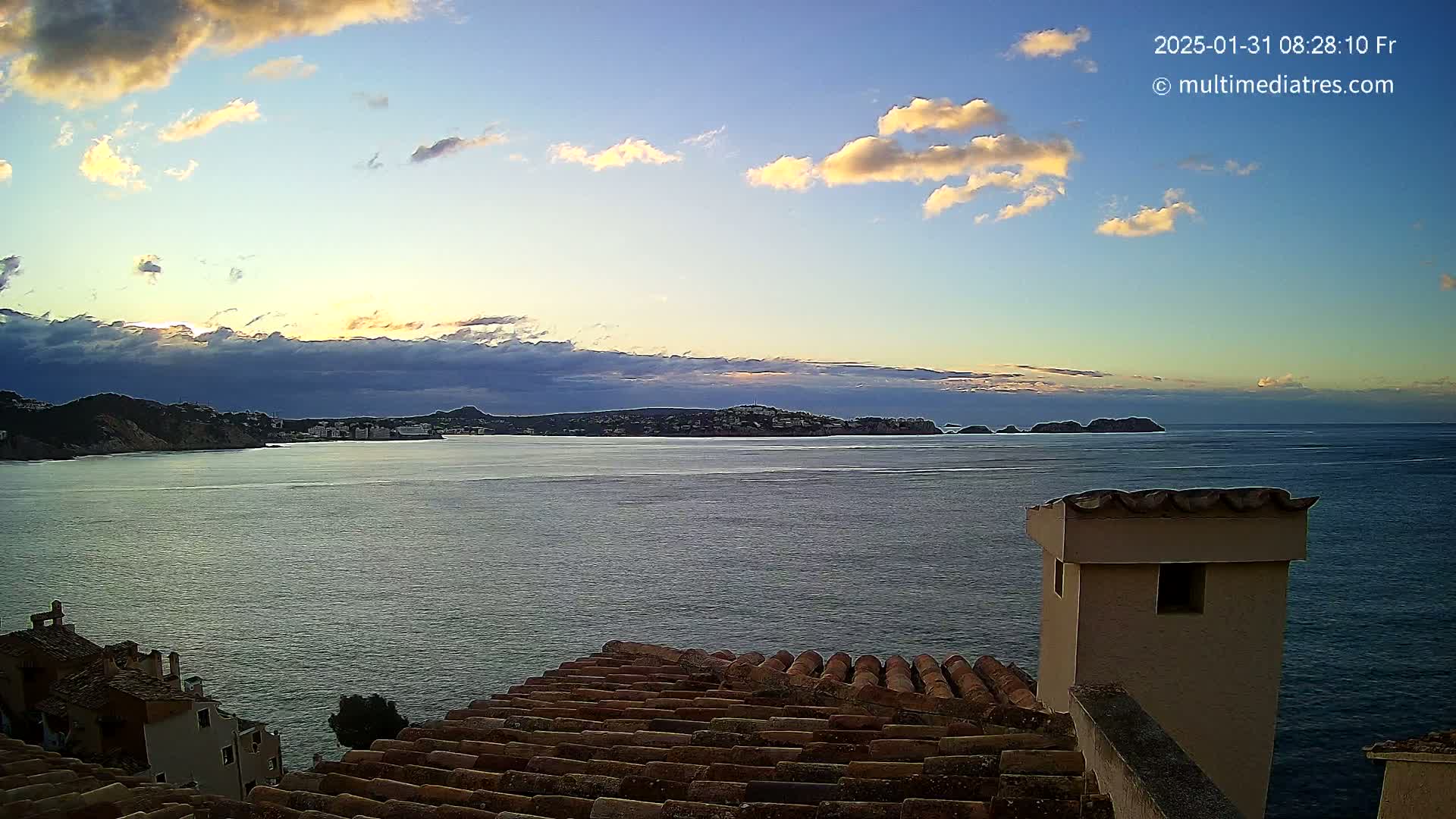 A calm sea at sunrise, partially cloudy, is viewed from a tiled roof, overlooking a town nestled against a coastal mountain range.