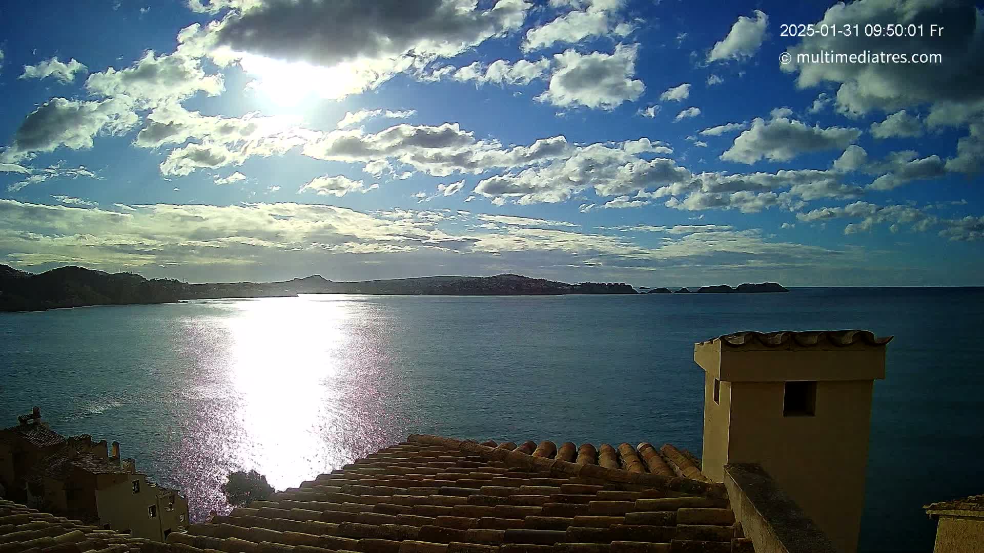 A partly sunny day with puffy clouds over a calm sea, viewed from a rooftop with terracotta tiles and a small building.