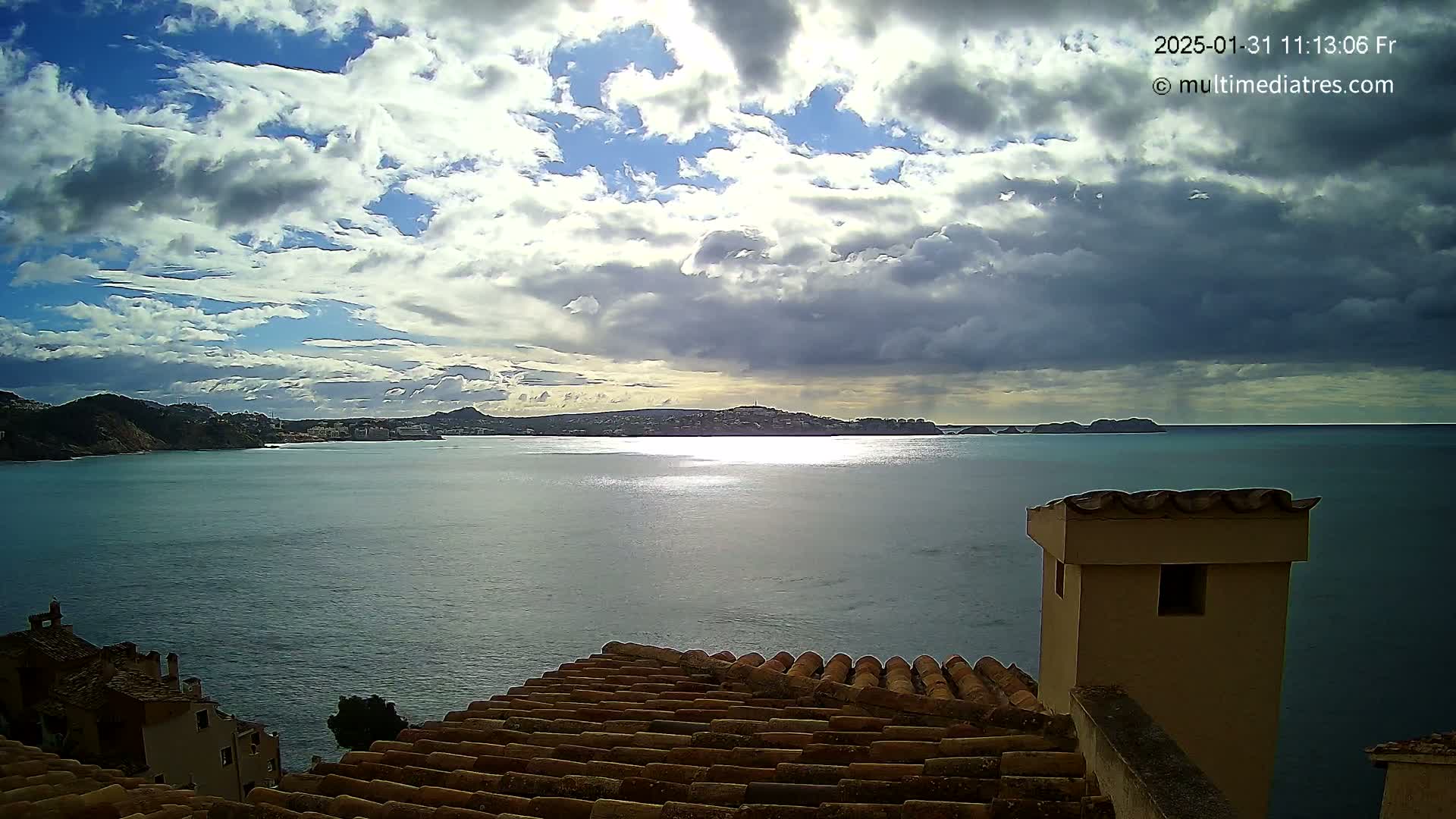A partly cloudy sky over a calm bay, viewed from a tiled rooftop with a chimney in the foreground.