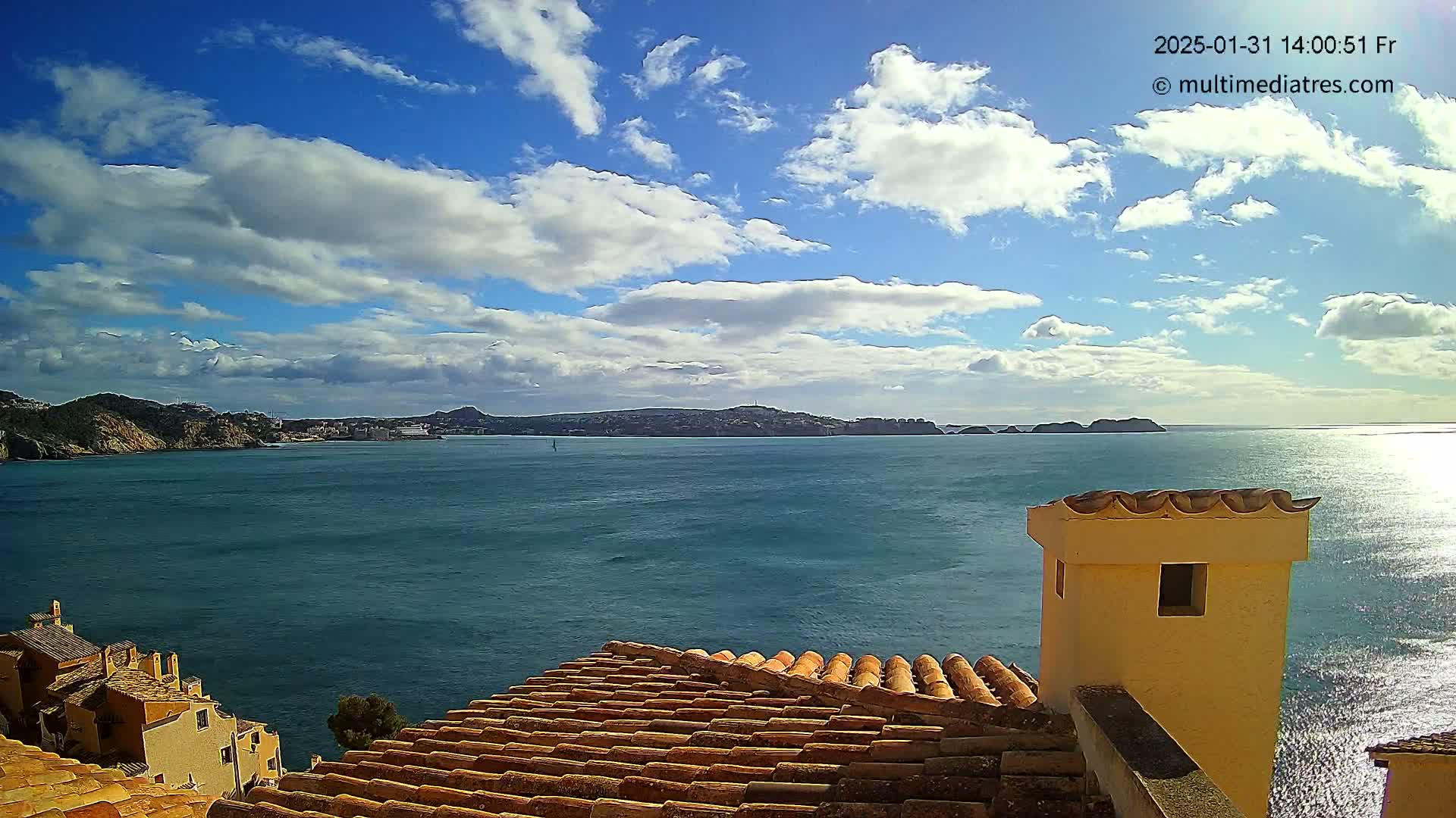 A partly cloudy sunny day overlooks a calm blue sea, with distant hills and a tiled rooftop in the foreground.
