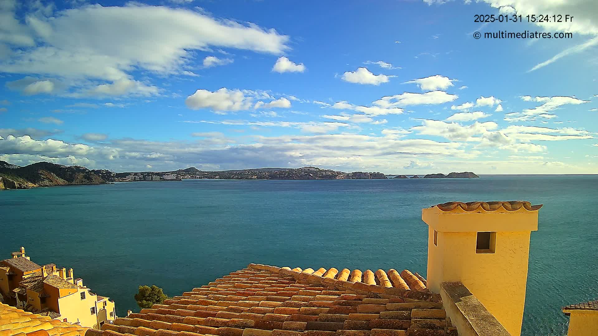 A partly cloudy blue sky overlooks a calm teal ocean, viewed from a terracotta roof with a pale yellow chimney in the foreground.