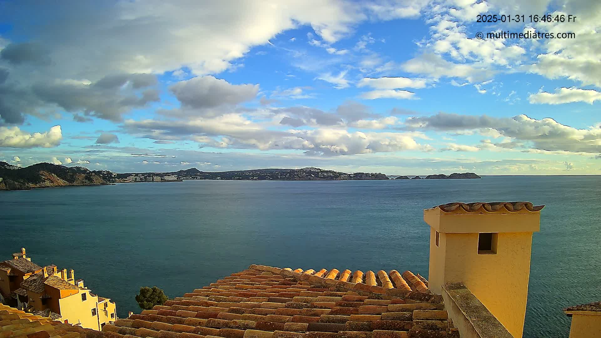 A partly cloudy sky overlooks a calm sea, with a coastal town and hills visible in the distance, seen from a vantage point of a terracotta roof and a small building's chimney.