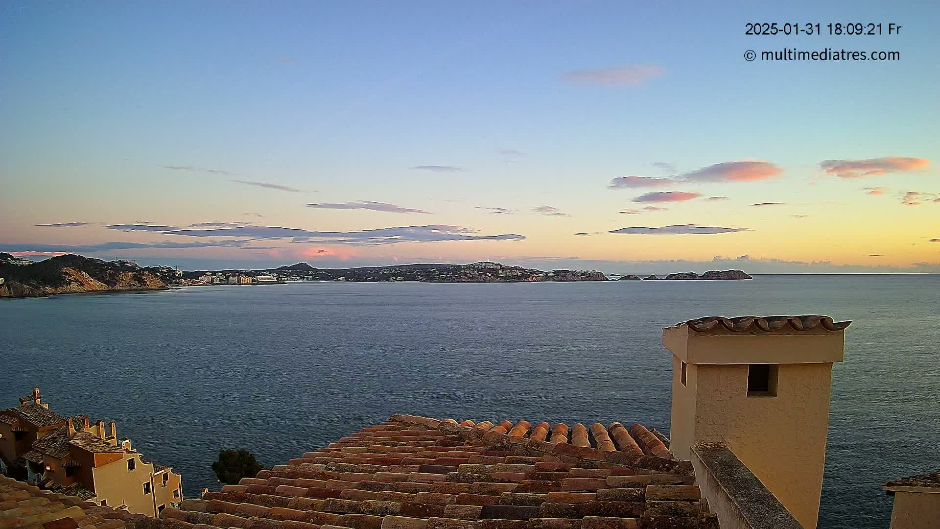A coastal scene at sunset shows a calm sea with a town and islands in the distance, viewed from a rooftop with terracotta tiles.