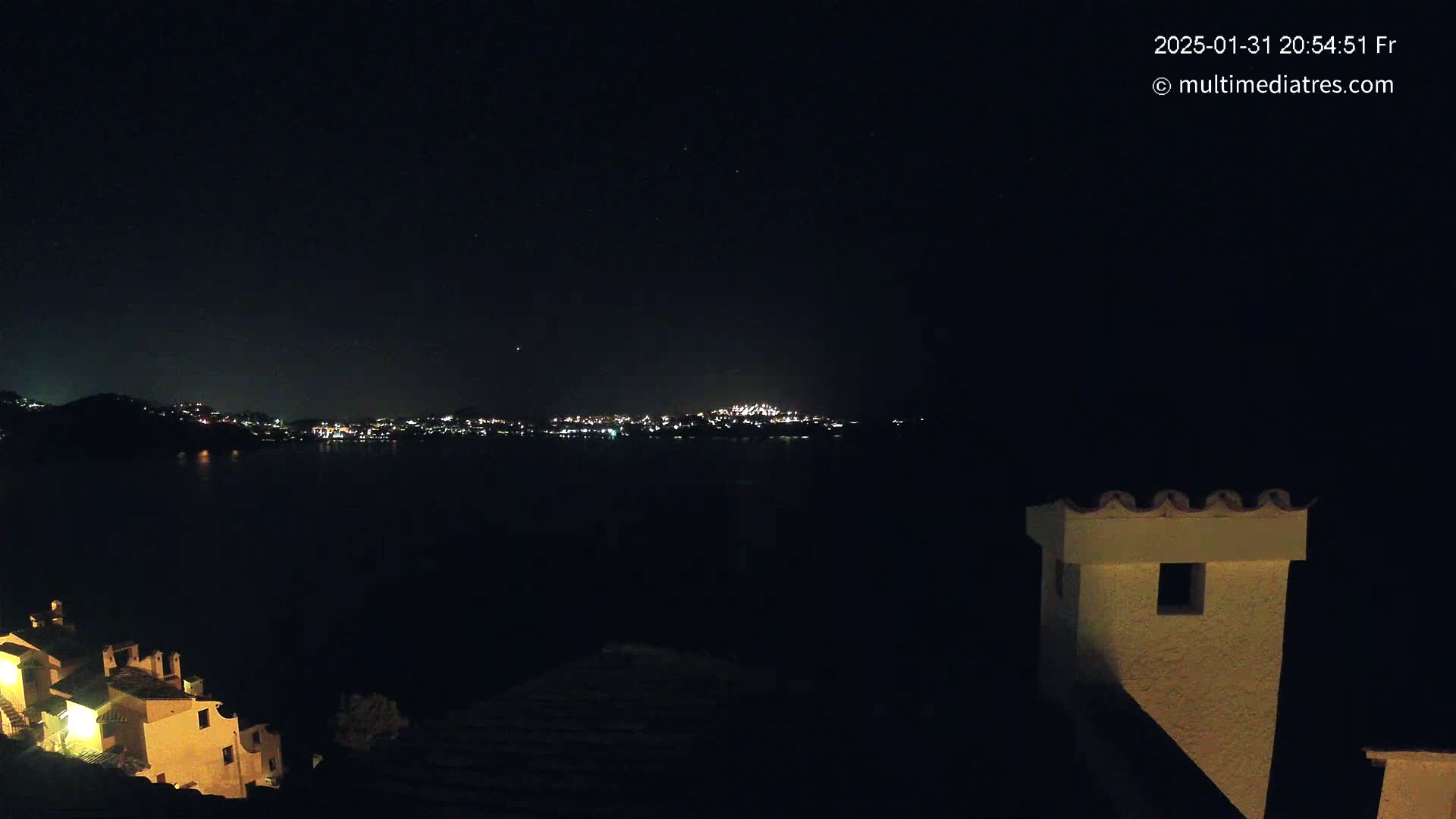 A nighttime view of a town's lights across a dark body of water, seen from a hilltop with a building's chimney in the foreground under a clear, starry sky.