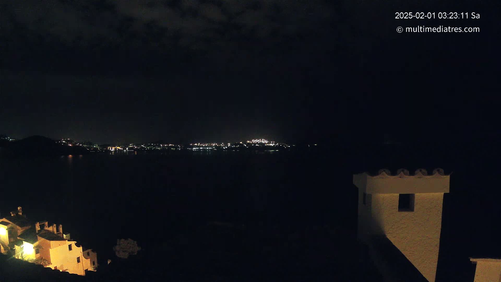 A nighttime view of a city skyline across a body of water, with a partially lit building in the foreground under a cloudy sky.
