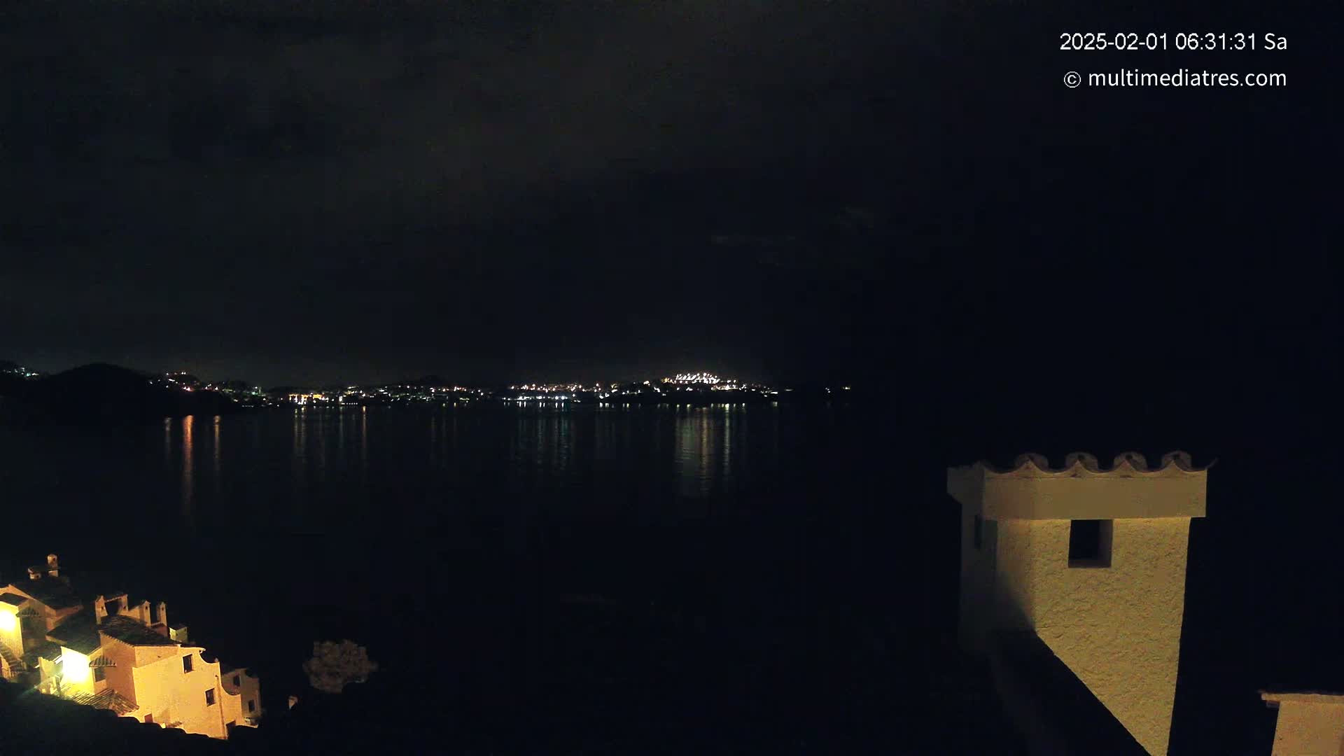 A nighttime view of a calm body of water reflecting the lights of a distant city, with a portion of a building visible in the foreground under a dark sky.