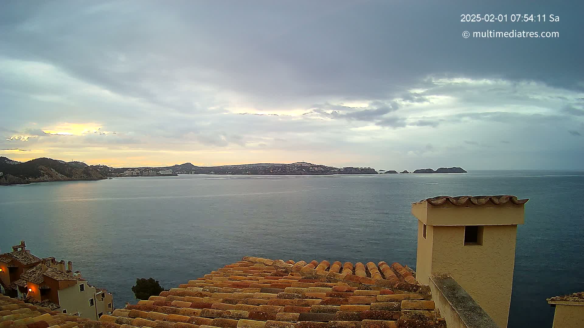 A partly cloudy sunrise over a calm sea, viewed from a tiled roof with a chimney in the foreground.