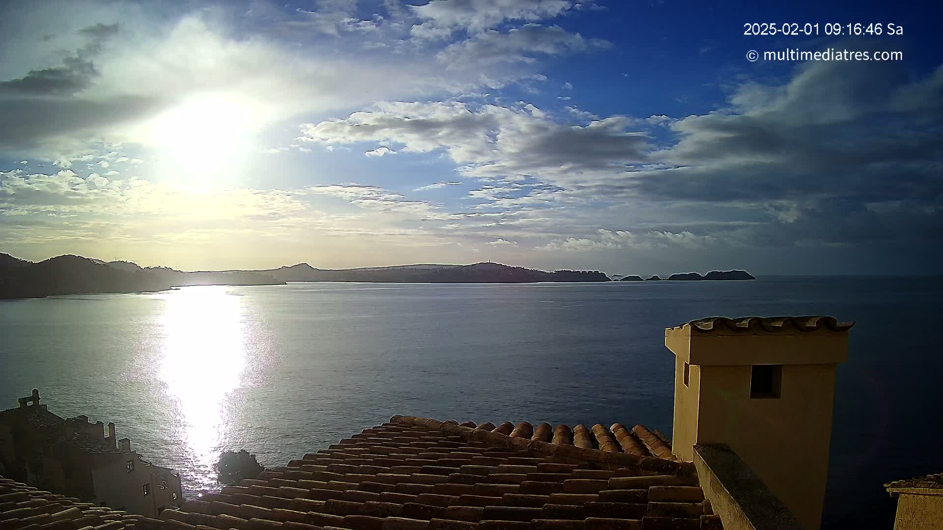 A partly cloudy sunrise over a calm sea, viewed from a tiled rooftop with a small building in the foreground.