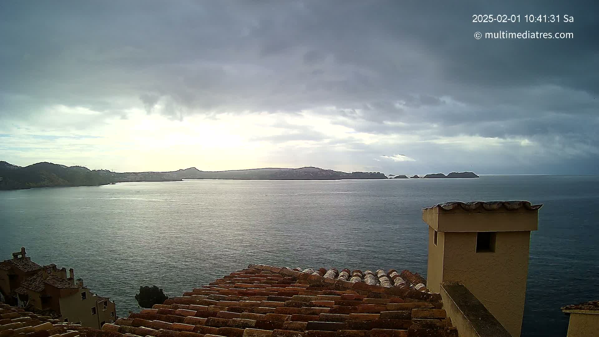 Overcast skies loom over a calm sea separating a coastal town with red-tiled rooftops from distant islands.