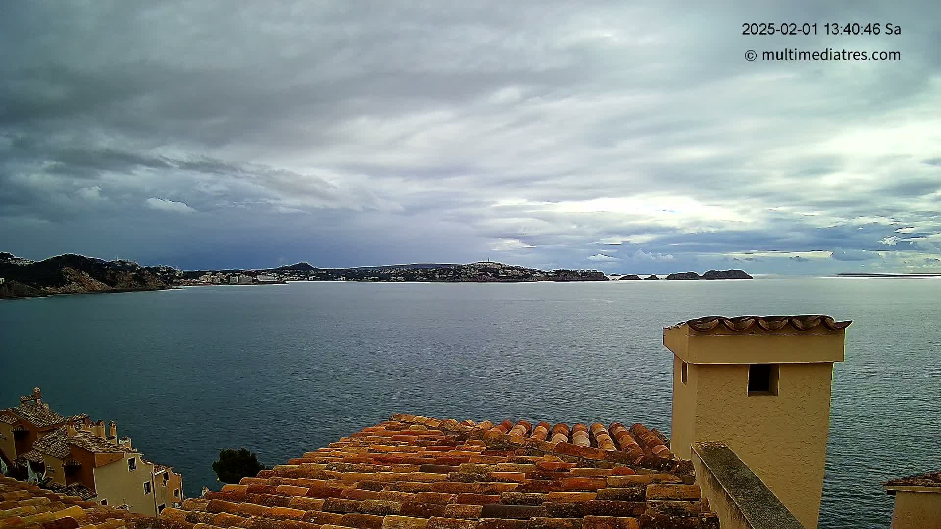 A view from a tiled rooftop shows a calm sea under a cloudy sky, with a distant town nestled between hills and small islands on the horizon.