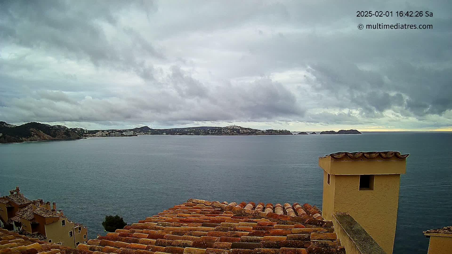 A view from a tiled rooftop shows a calm sea under a cloudy sky, with a distant coastline and buildings visible across the water.