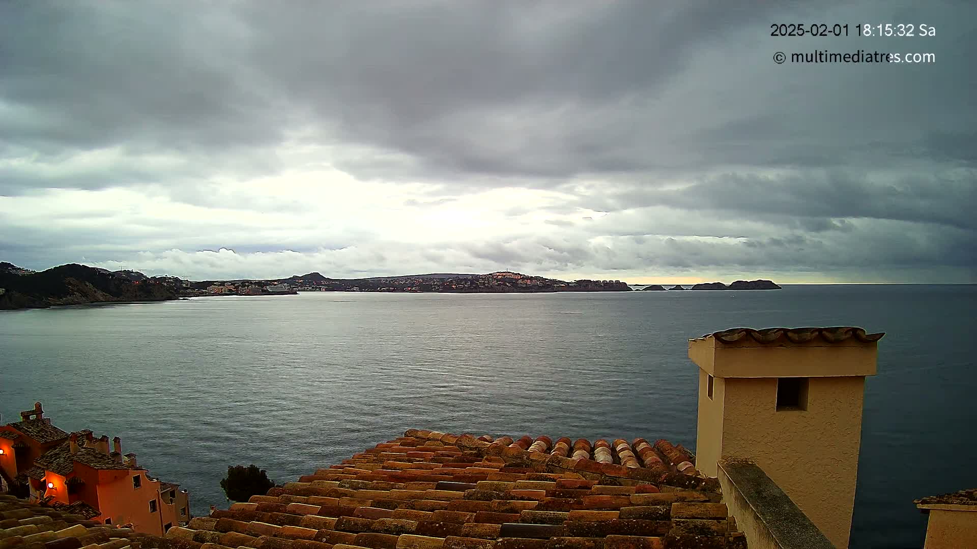 A coastal town is visible across a calm body of water under a cloudy sky, viewed from a rooftop with terracotta tiles.