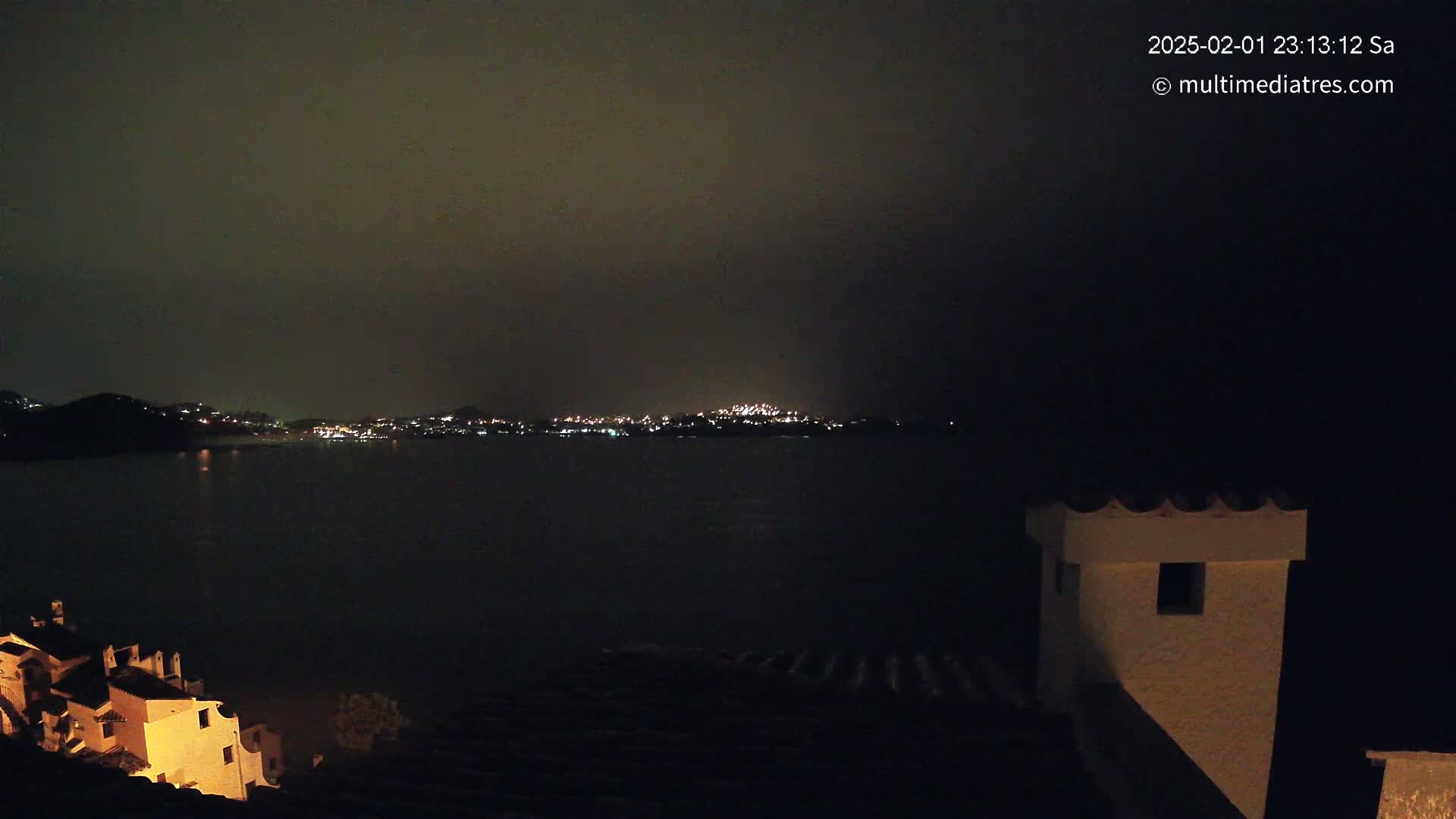 A nighttime view of a dark body of water with a city skyline faintly lit on the far shore, seen from a hilltop with buildings partially visible in the foreground under a dark, possibly overcast sky.