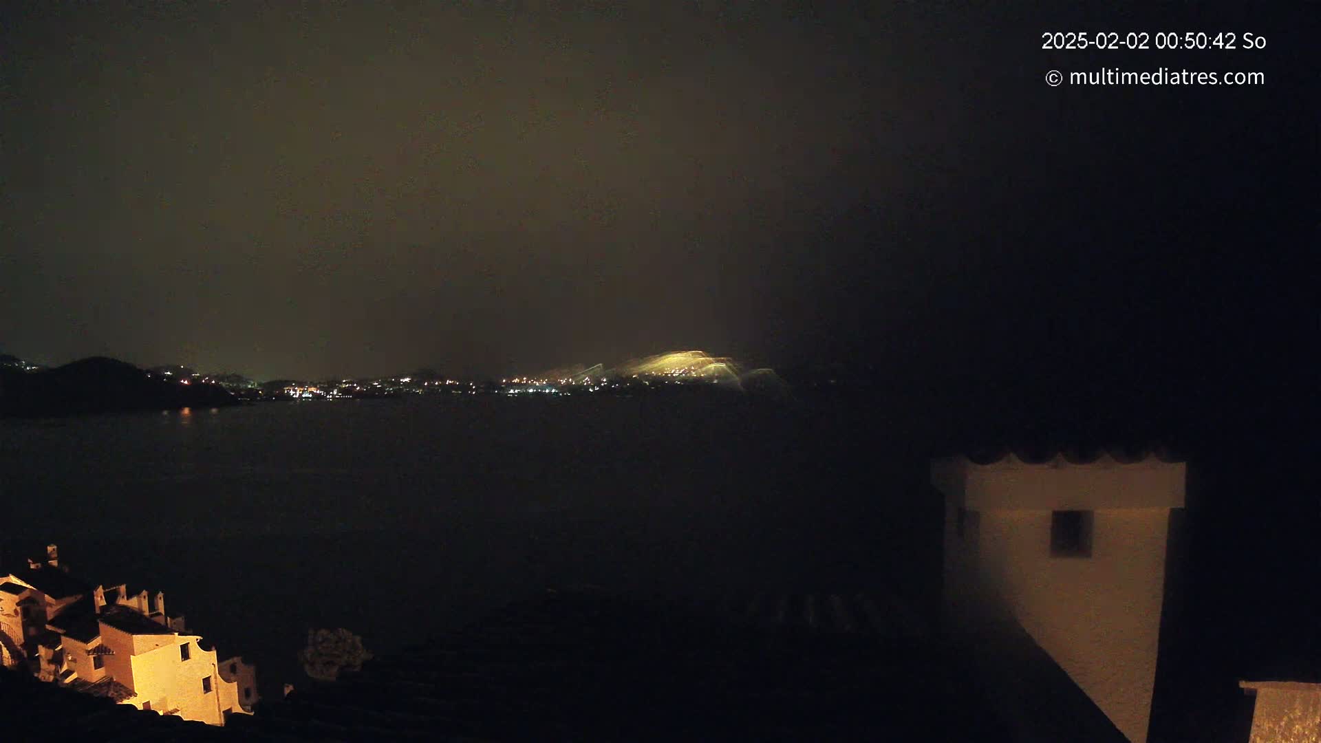 A nighttime view across a dark body of water shows a dimly lit town on the opposite shore, with a hilltop glowing brightly, and buildings illuminated from below on the near shore.