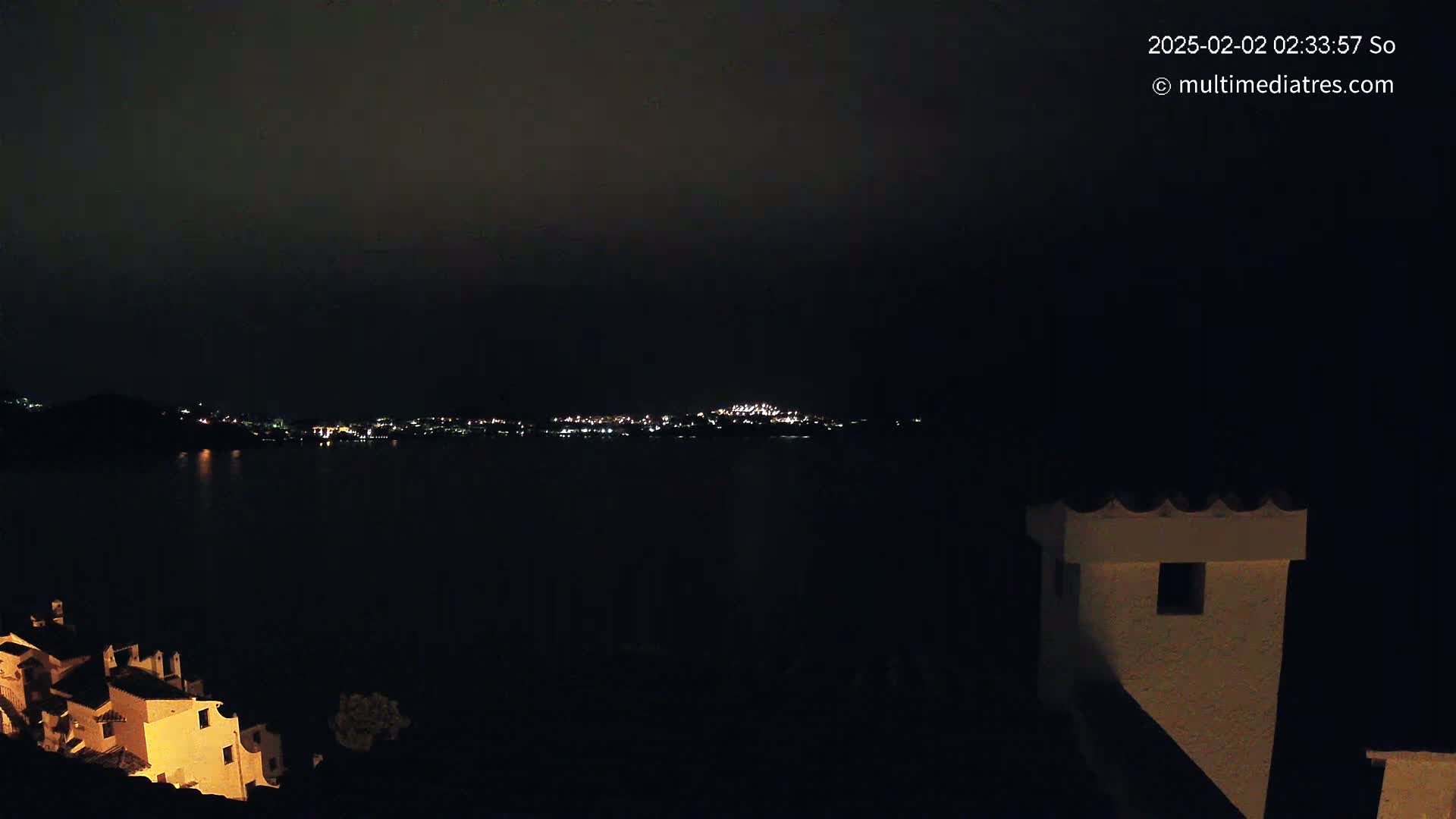A nighttime view of a city across a dark body of water, with illuminated buildings visible in the foreground and background under a cloudy sky.