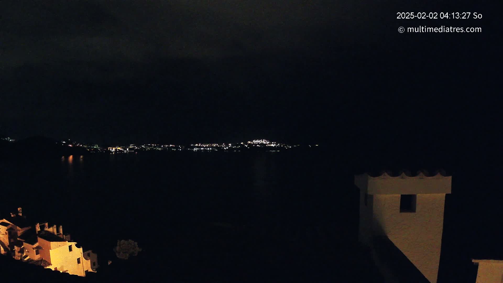 A nighttime view shows a dark body of water reflecting the lights of a distant town, with illuminated buildings in the foreground under a cloudy sky.