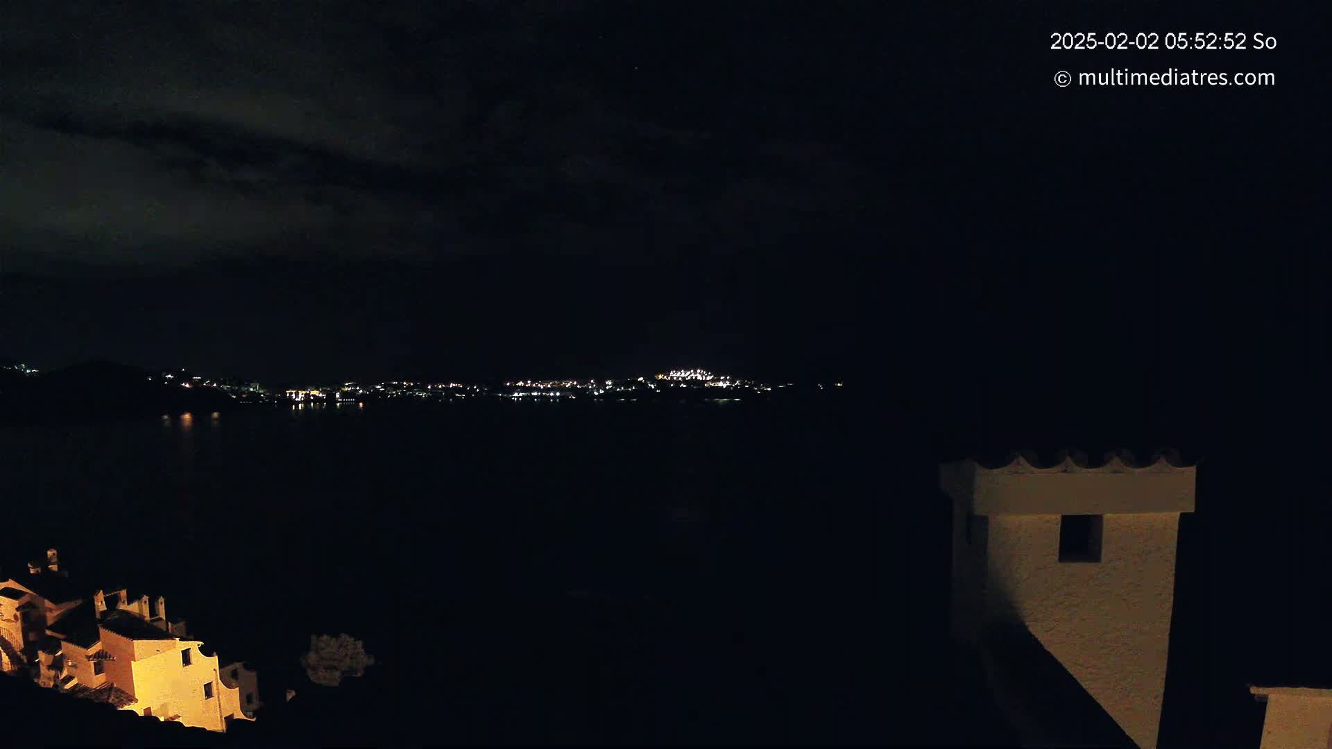 A nighttime view shows a dark body of water reflecting the lights of a distant city across the water, with a portion of a building and its chimney visible in the foreground under a cloudy sky.