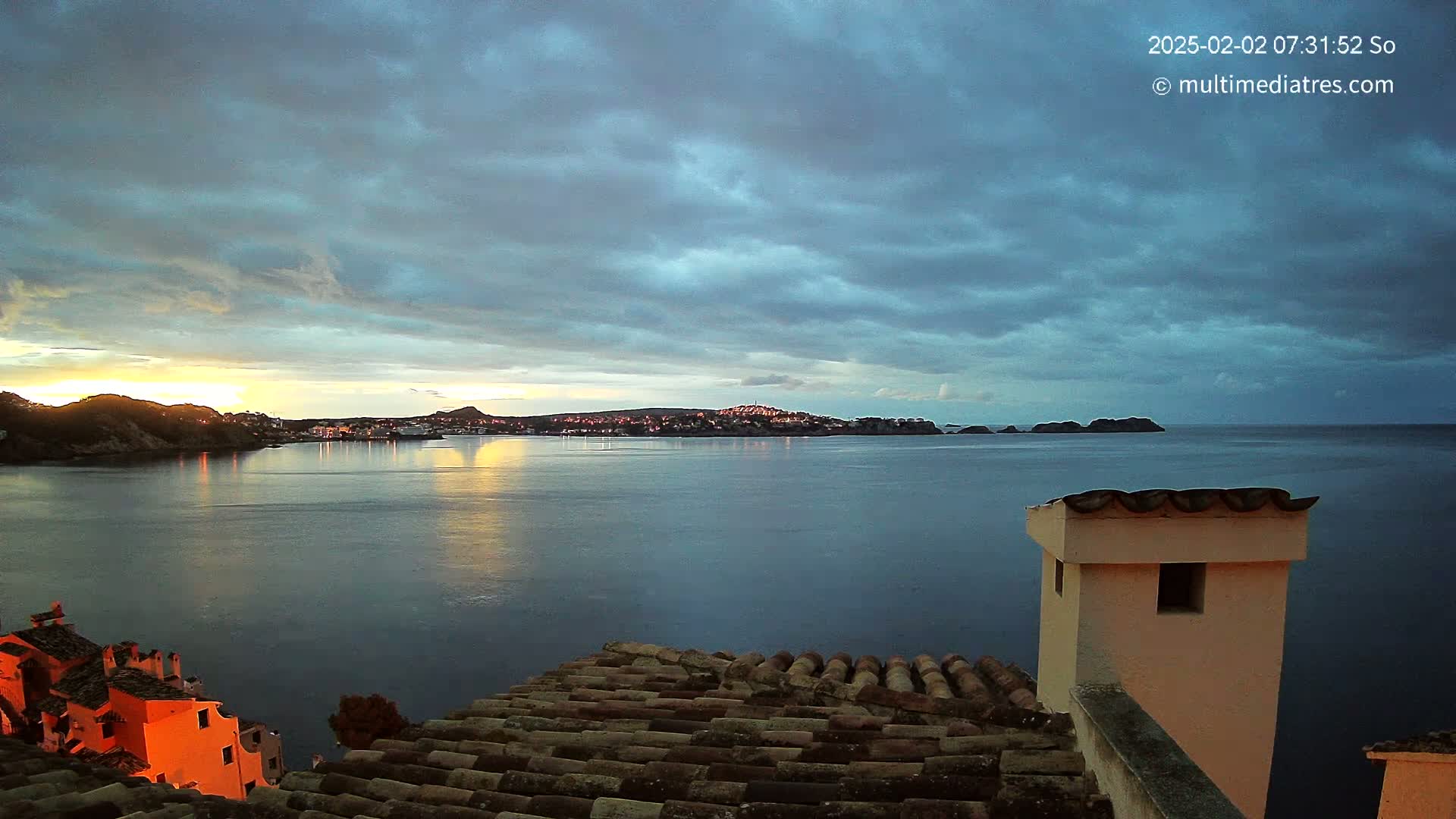 A coastal town at sunrise is viewed from a rooftop, with a calm sea reflecting the cloudy sky.