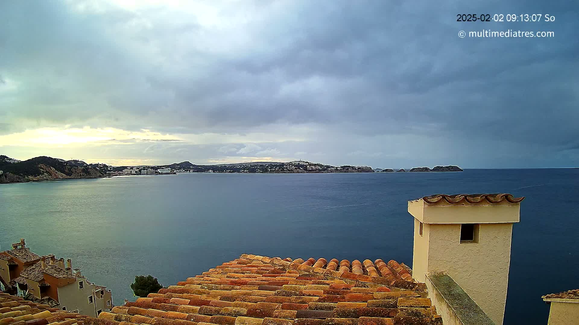 A coastal town is seen from a rooftop, under a cloudy sky, with a calm sea stretching to a distant hilly shoreline.