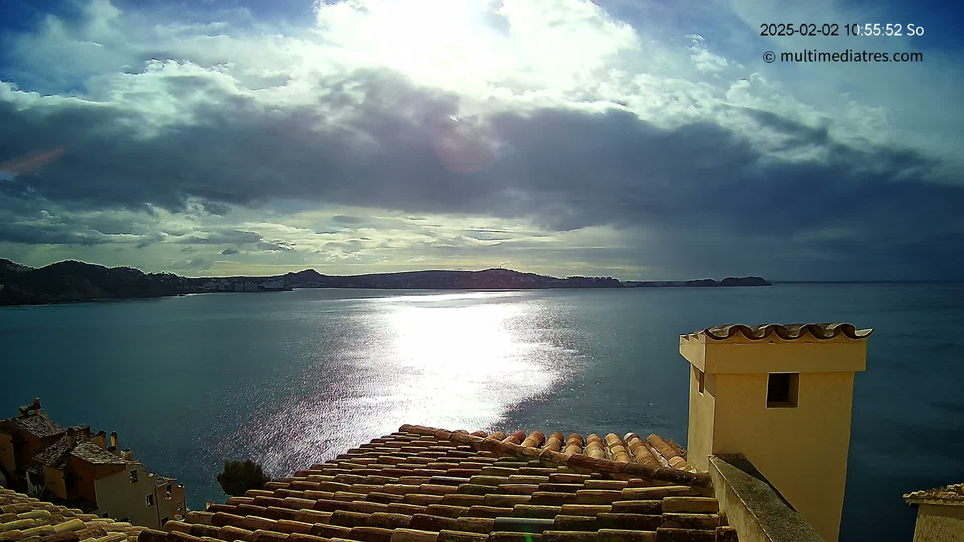 A partly cloudy sky over a calm sea, reflecting sunlight, is viewed from a tiled rooftop, near a building with a light-yellow chimney.