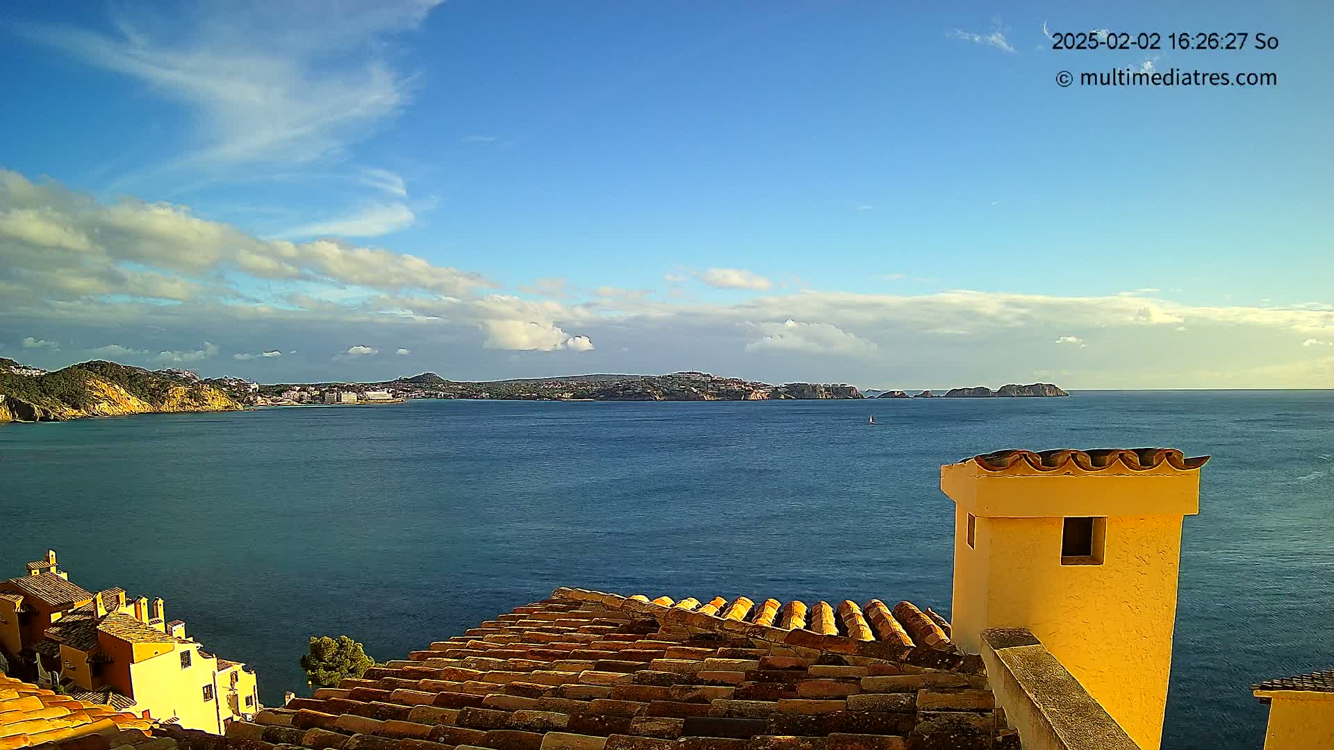 A partly cloudy blue sky overlooks a calm blue sea, with a distant coastline and a portion of a terracotta roof and yellow chimney in the foreground.