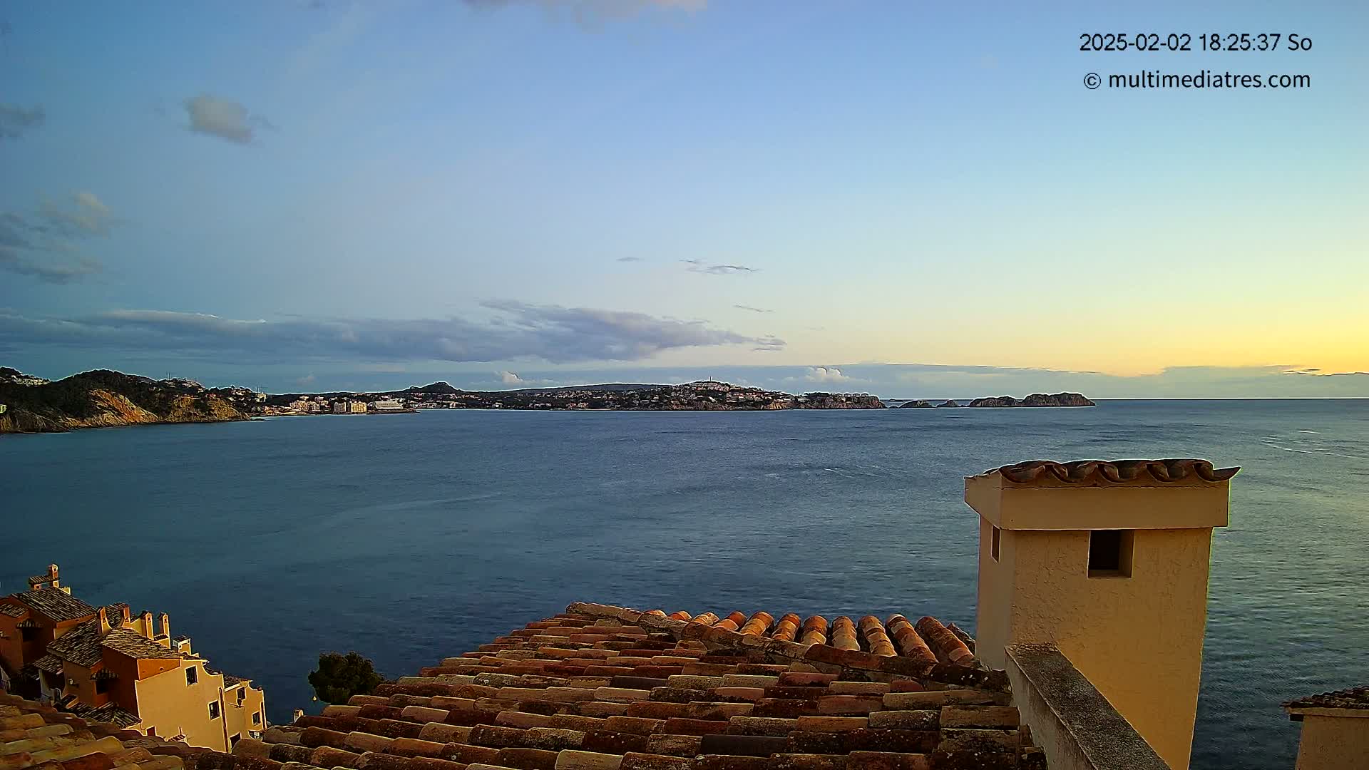 A tranquil coastal scene at sunset, featuring a calm sea, distant hills with buildings, and a view from a tiled rooftop.
