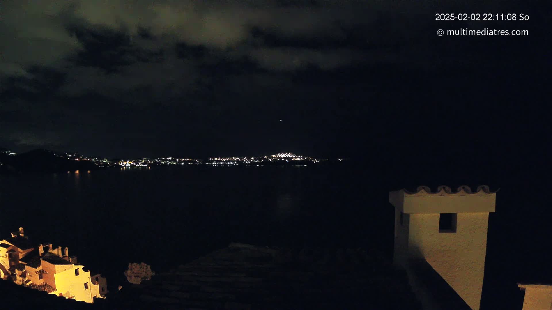 A nighttime view of a city's lights across a dark body of water, under a cloudy sky, is seen from a vantage point near a building with a visible chimney.