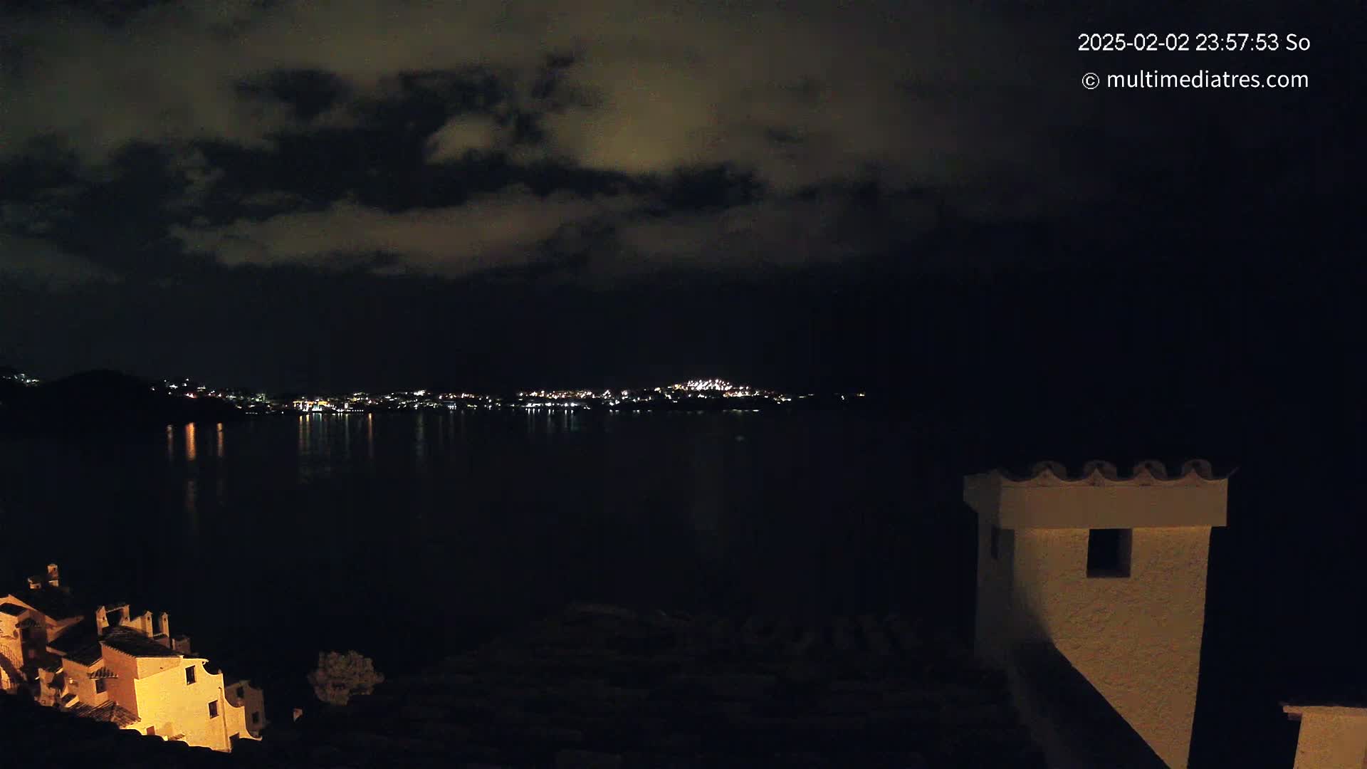 A nighttime view of a city across a dark body of water, under a cloudy sky, with illuminated buildings visible in the foreground.