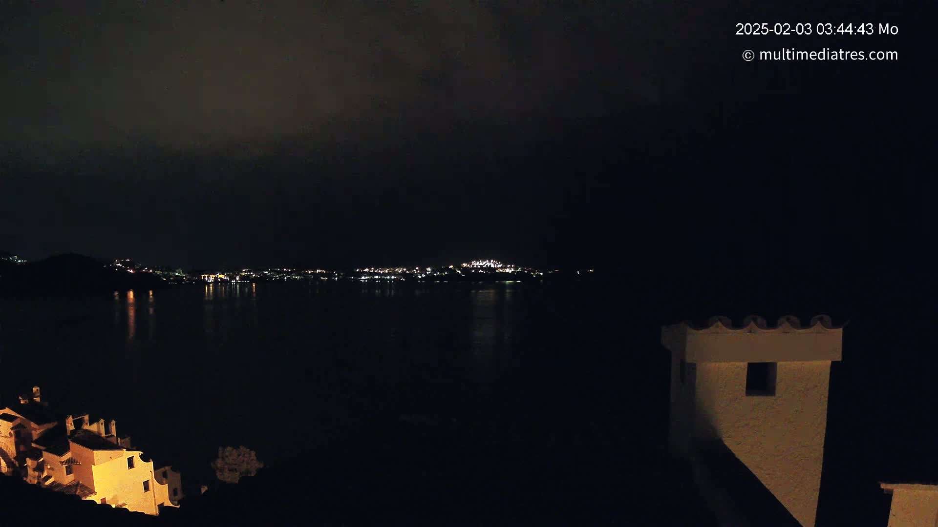 A nighttime view shows a dark body of water reflecting the lights of a city on the opposite shore, with a building and its chimney visible in the foreground under a cloudy sky.