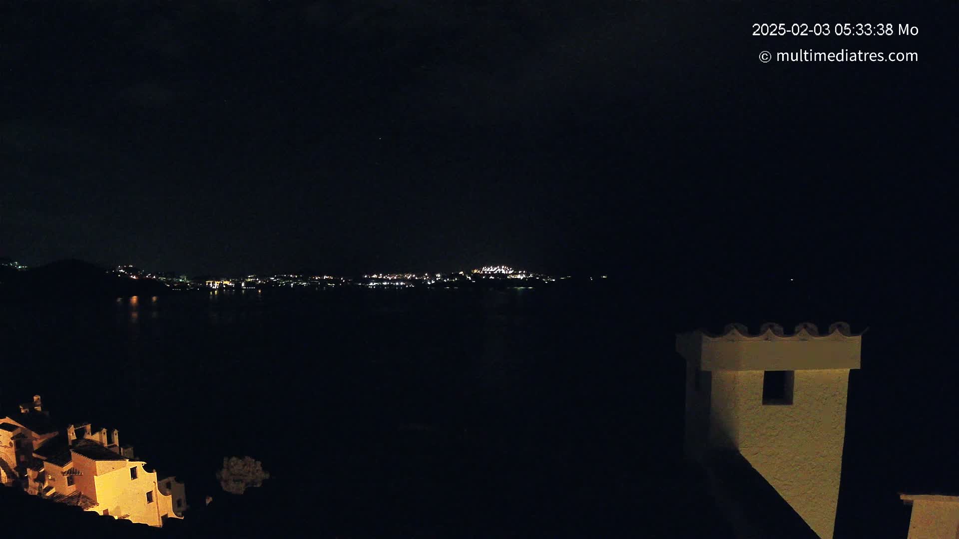 A nighttime view shows a dark body of water separating a dimly lit shoreline from a brightly lit town across the water, with a portion of a building and its chimney visible in the foreground under a clear, dark sky.