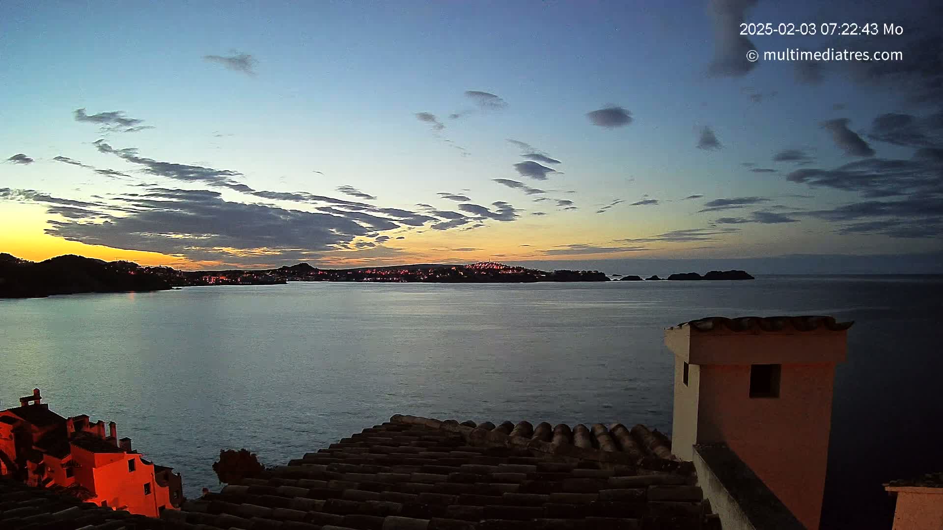 A coastal town at twilight is seen from a rooftop, with a calm sea and a partly cloudy sky showing the last colors of sunset.
