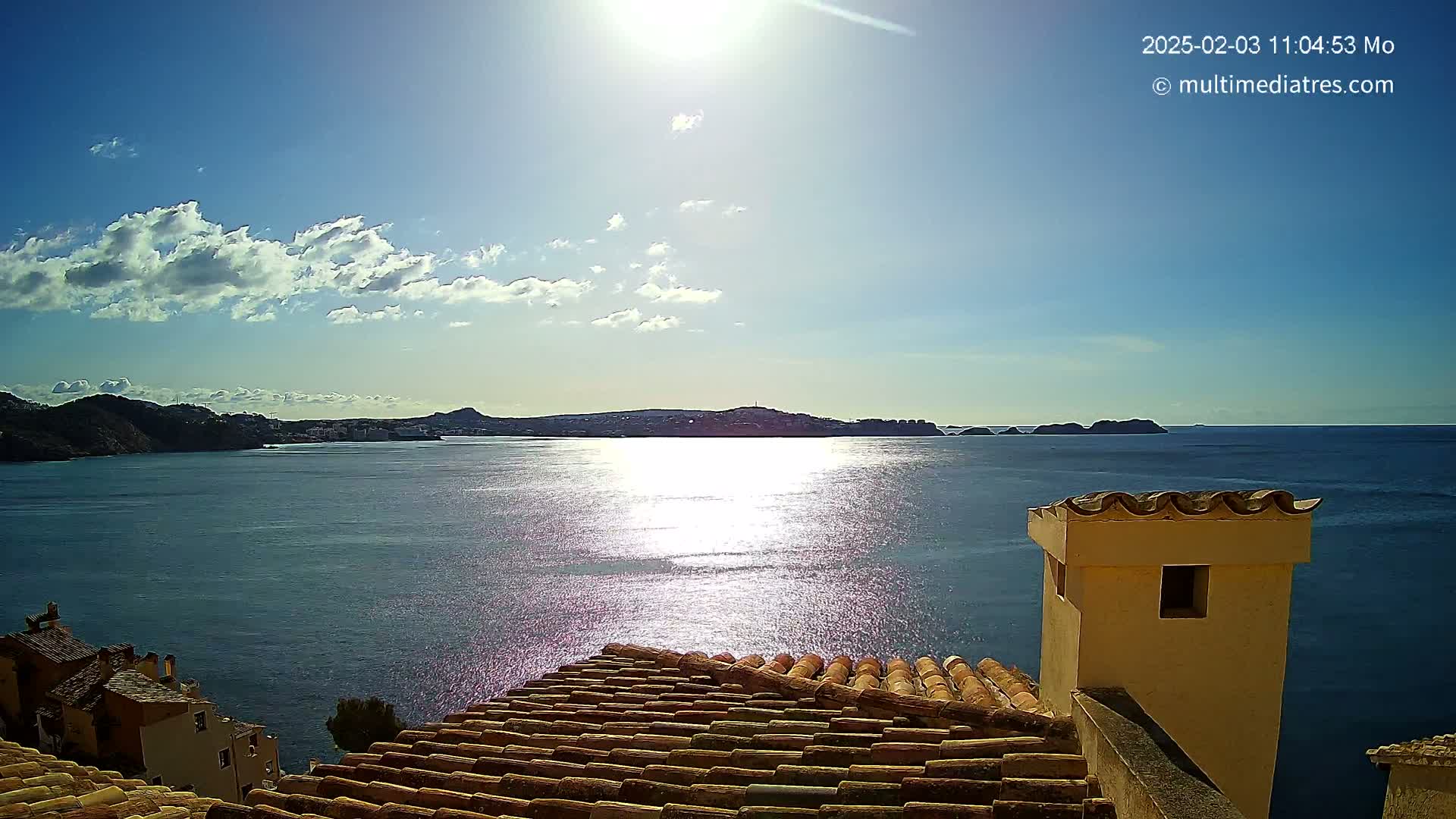 A partly sunny day reveals a sparkling blue sea, with distant hills and islands under a bright sky, as seen from a tiled rooftop with a chimney.