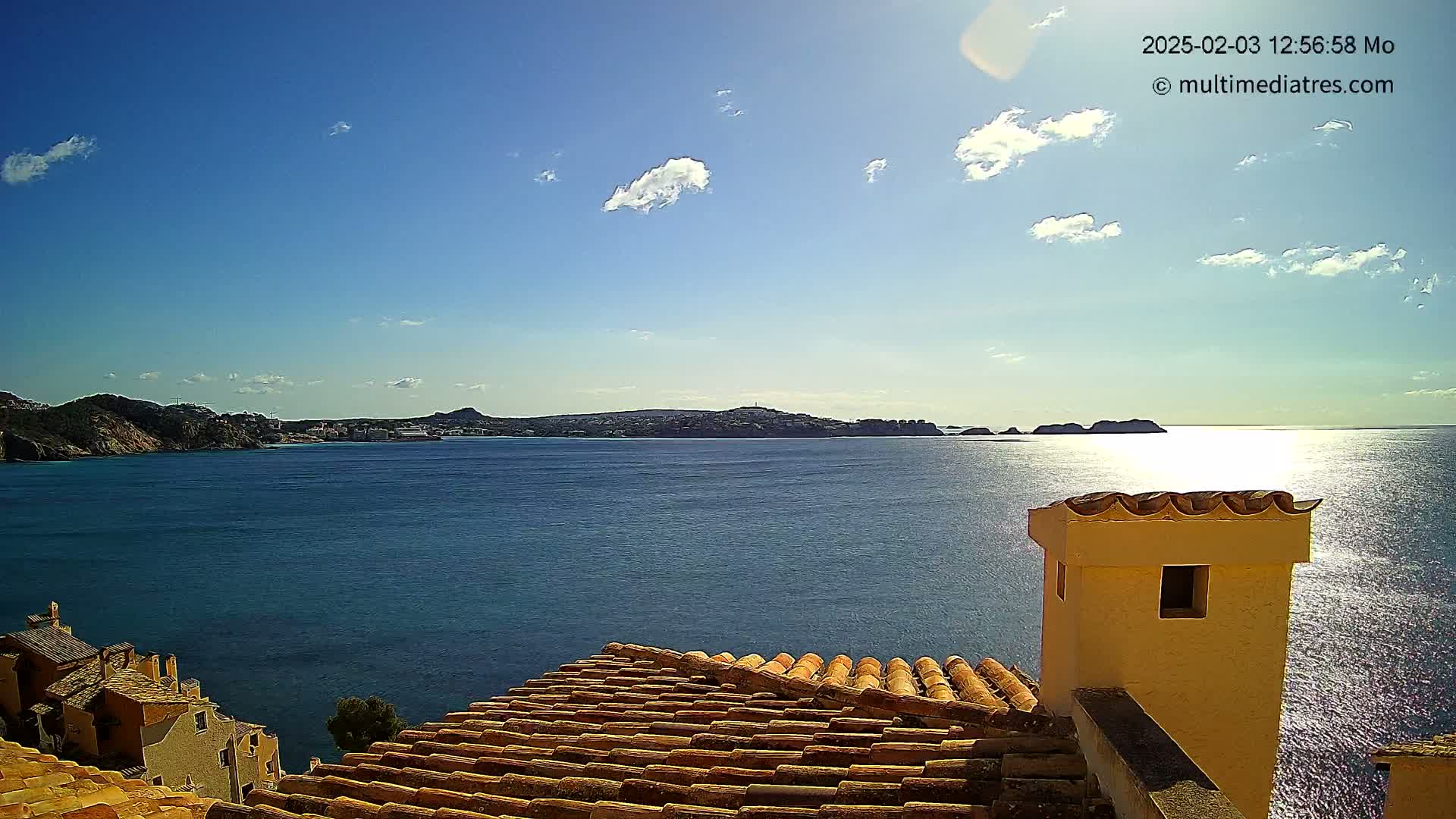 A partly sunny day overlooks a calm, blue sea with a distant coastline, viewed from a terracotta roof with a pale yellow chimney.