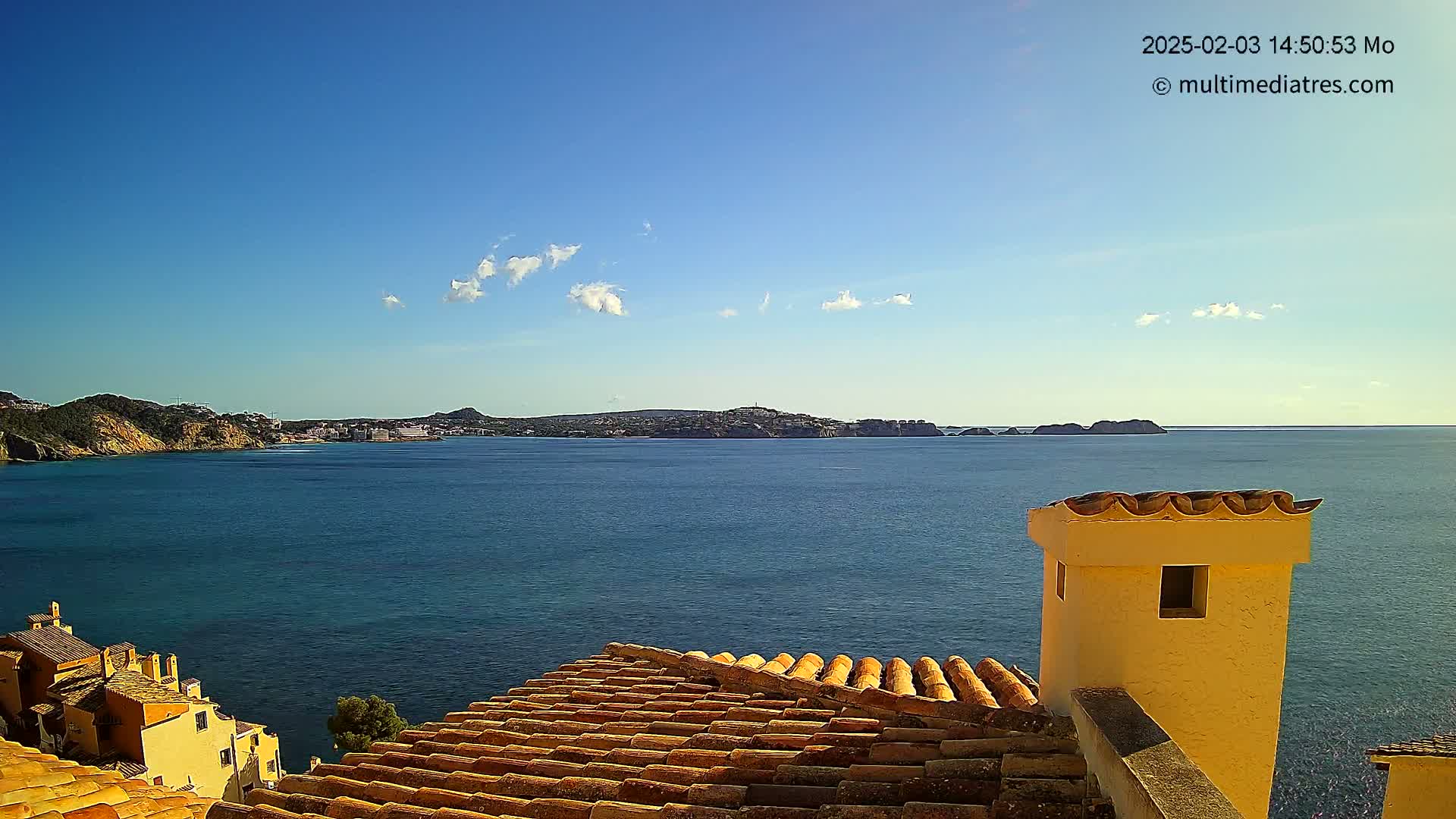 A coastal view on a sunny day shows a calm blue sea, distant islands, and a portion of a terracotta roof with a yellow chimney in the foreground.