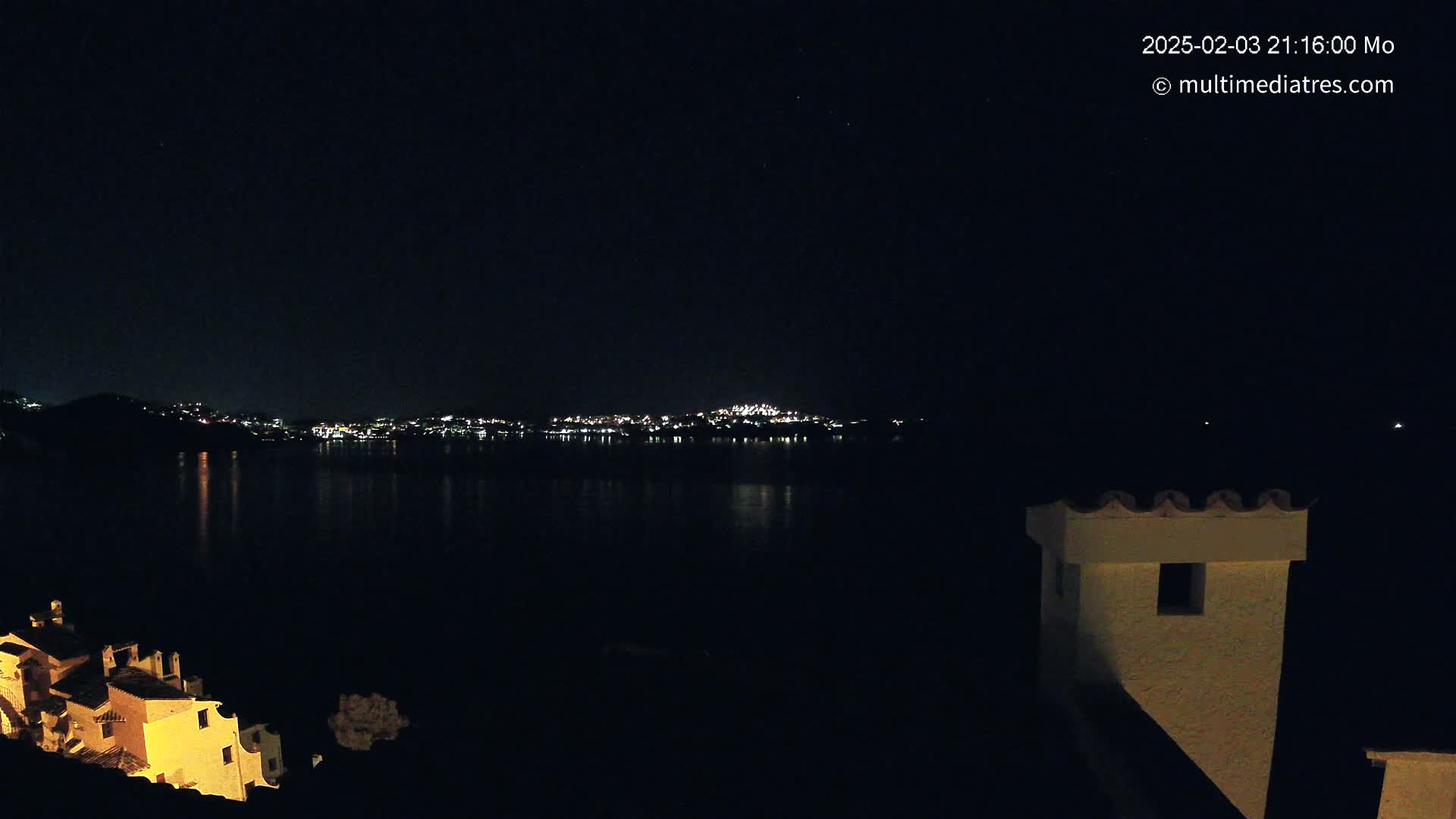 A nighttime view across a dark body of water shows a distant city skyline illuminated by lights, with a partially visible building in the foreground under a clear, starlit sky.
