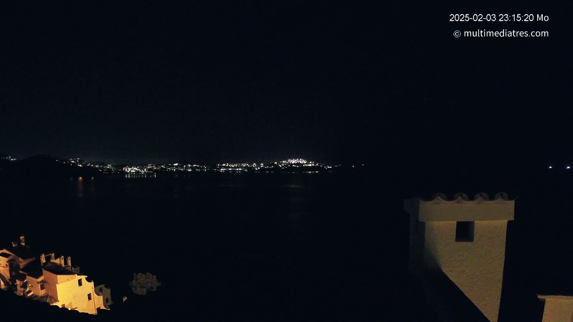 A nighttime view of a city's lights across a dark body of water, partially obscured by a building's silhouette in the foreground under a clear, star-filled sky.