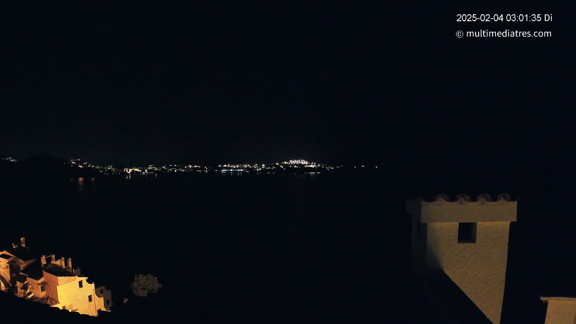 A nighttime view of a city skyline across a body of water, partially obscured by darkness, with a building's illuminated chimney in the foreground.