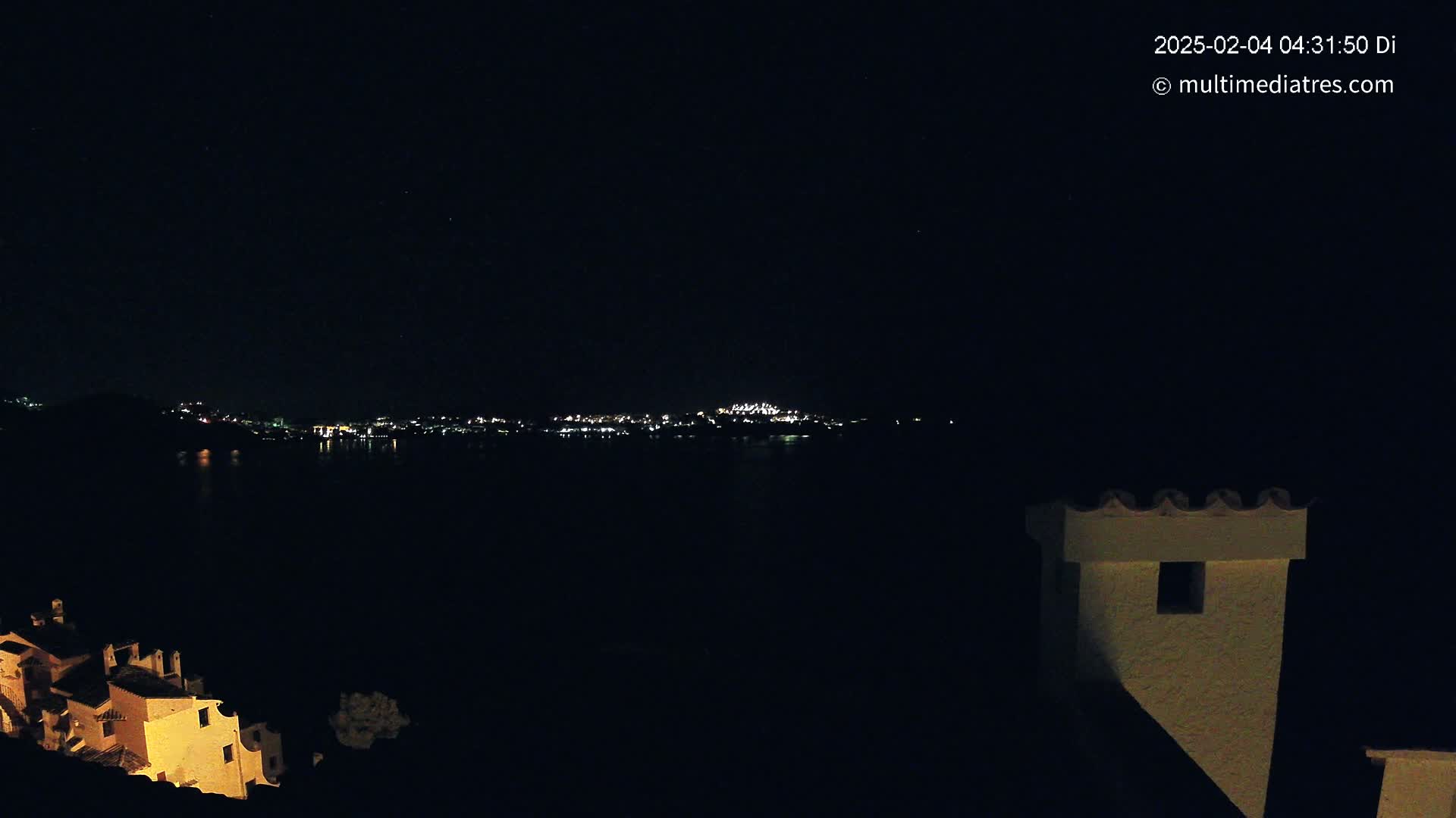 A nighttime view of a town's lights across a dark body of water, seen from a building's rooftop under a clear, starry sky.