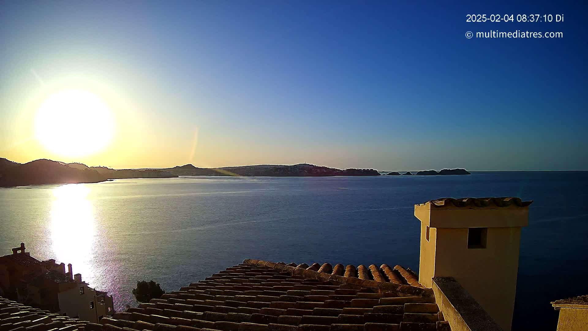 A bright sunrise over a calm sea, viewed from a rooftop with terracotta tiles and a chimney.