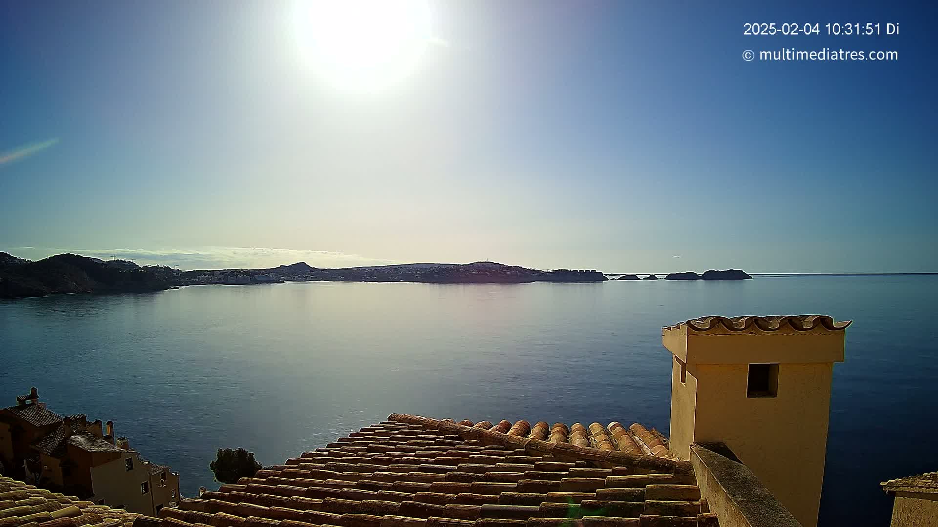 A bright, sunny day overlooks a calm body of water from a tiled rooftop, with a distant coastline visible on the horizon.