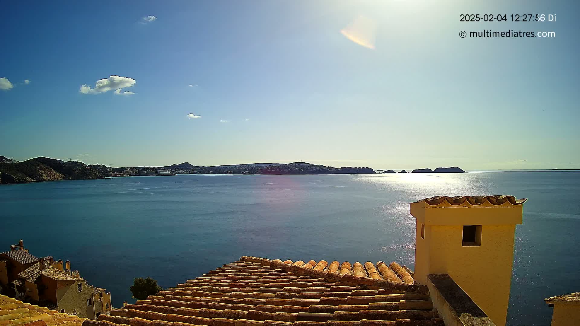 A sunny, cloudless day reveals a coastal town's terracotta rooftops overlooking a calm blue sea and distant land.