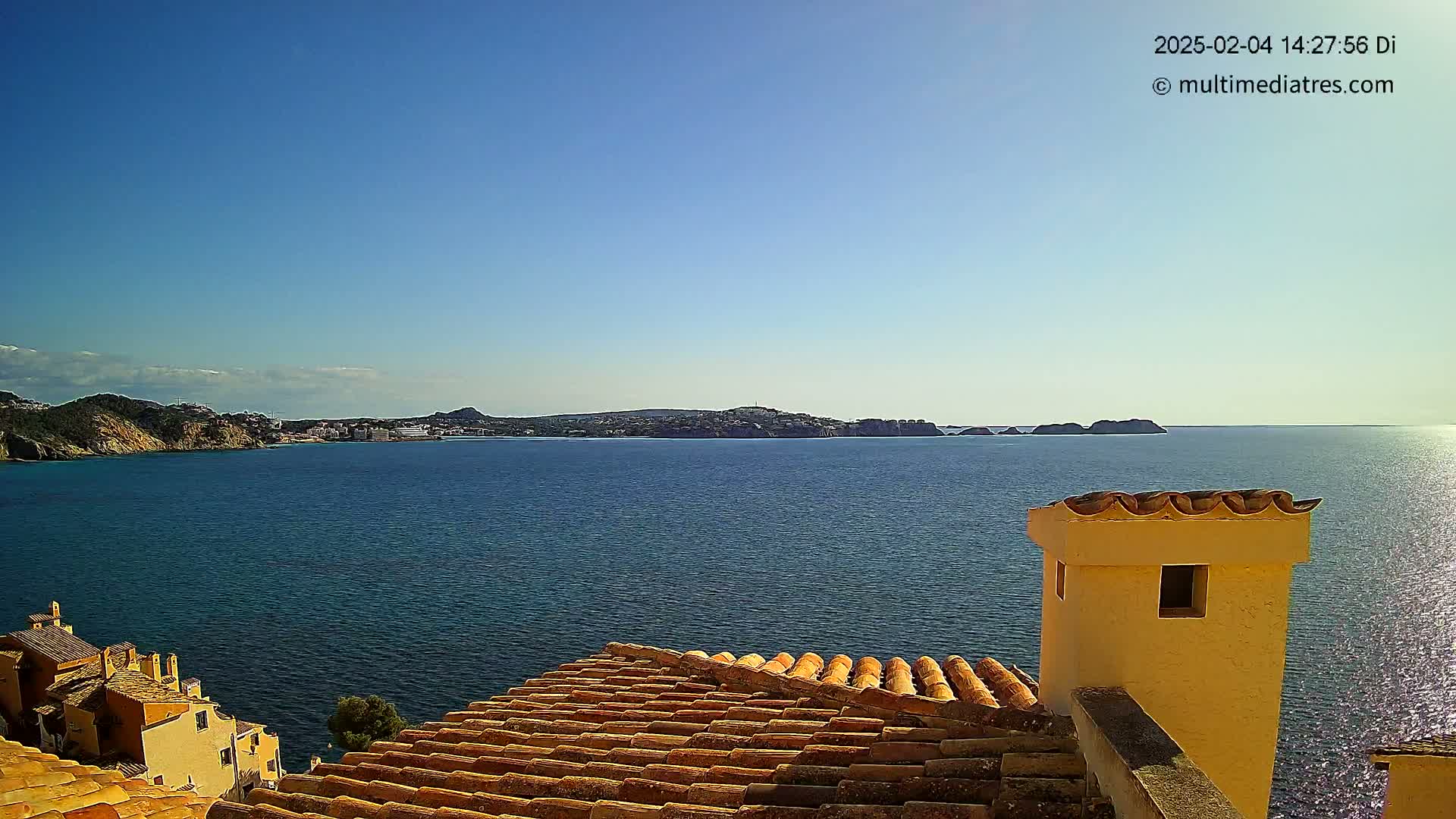 A coastal scene under a clear blue sky shows a calm sea, a distant town nestled against rocky hills, and a portion of a tiled roof with a stucco chimney in the foreground.