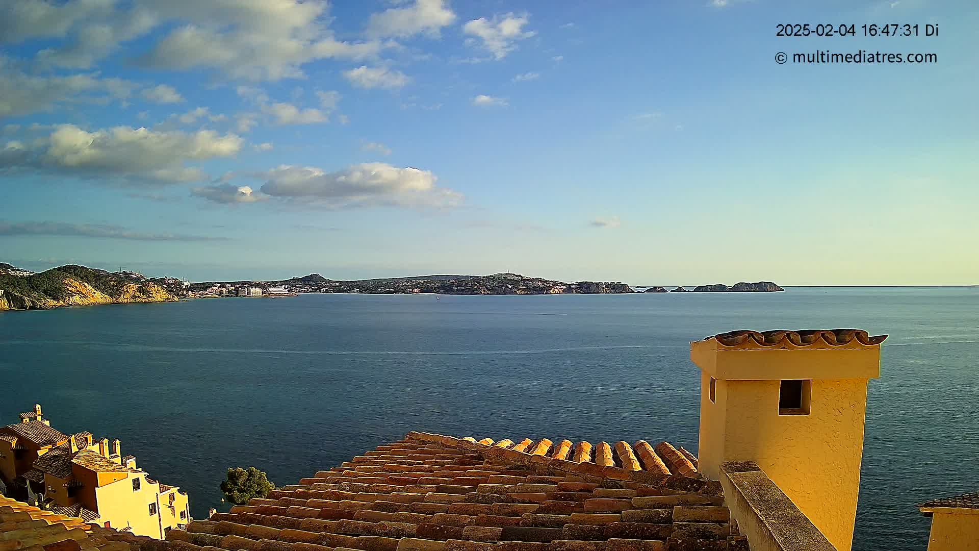 A partly cloudy day reveals a coastal town and tranquil blue sea, viewed from a tiled rooftop with a pale yellow chimney.