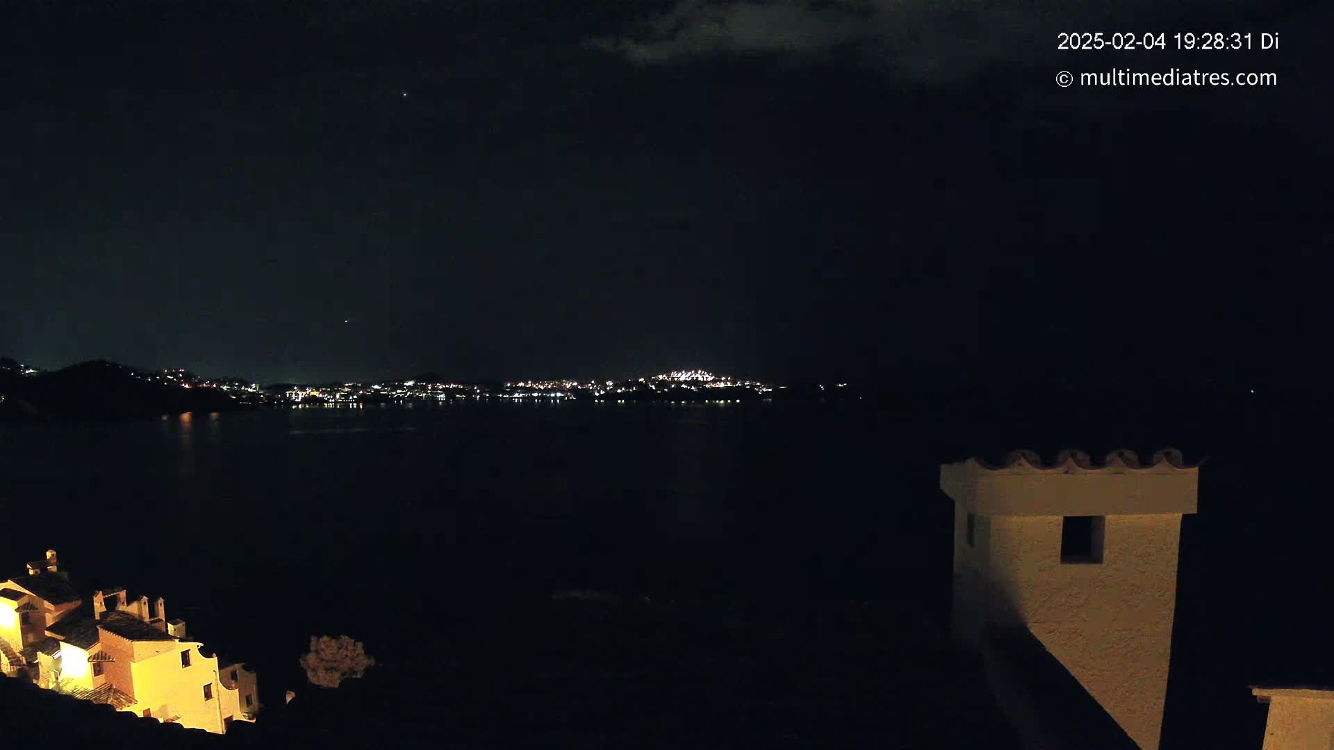 A nighttime view across dark water shows a distant city glowing with lights under a mostly clear, dark sky, with a partially lit building in the foreground.