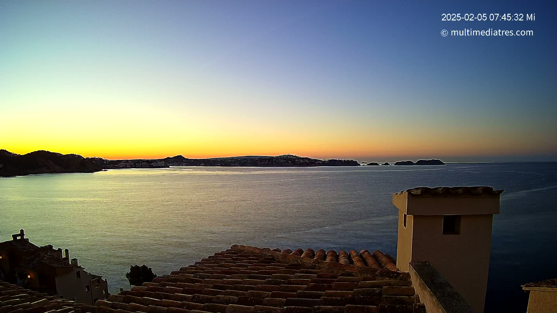 A tranquil sunrise over a calm sea, viewed from a rooftop with terracotta tiles, shows a coastal town nestled between hills.