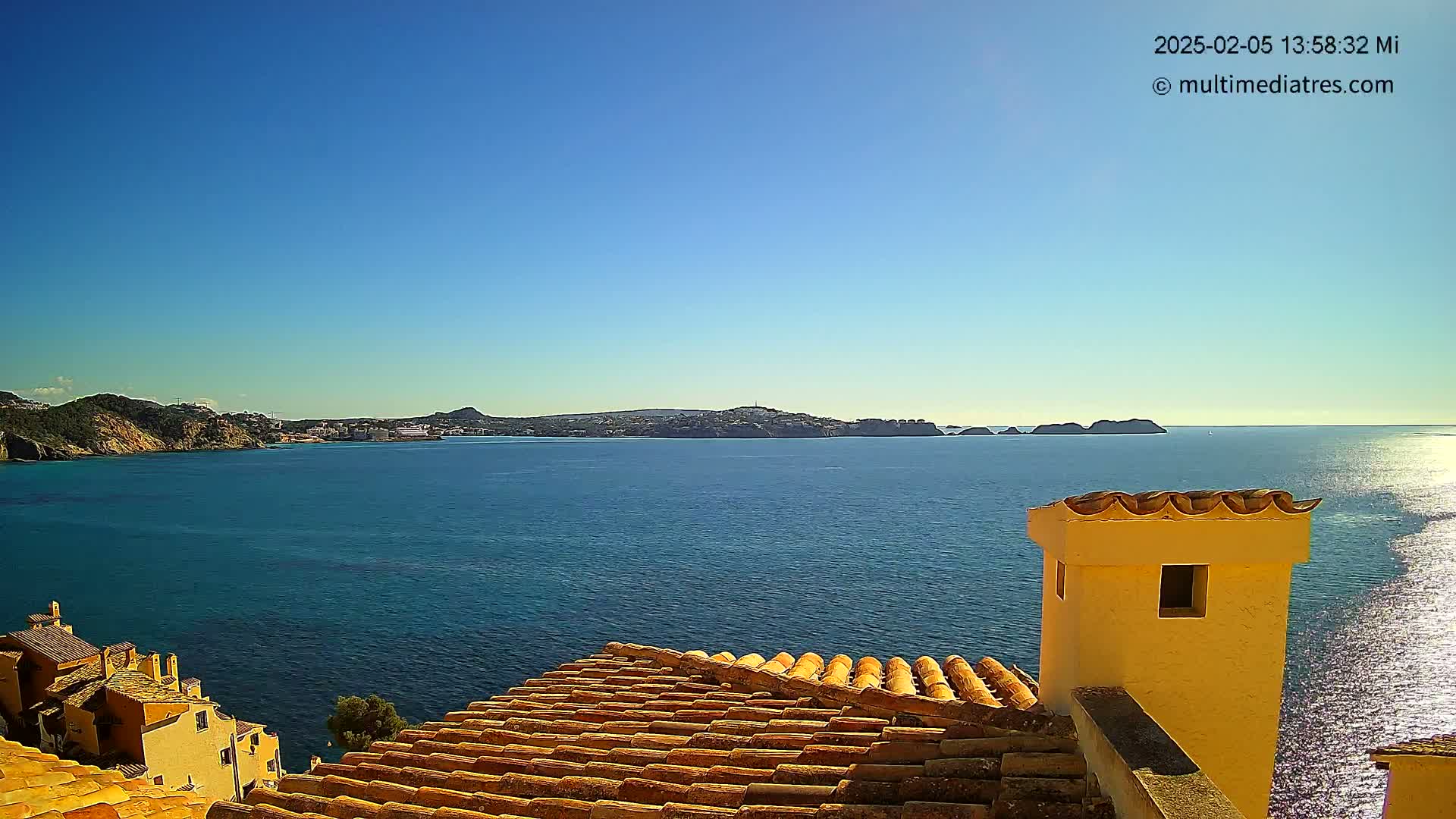 A terracotta roof and a yellow chimney overlook a calm, blue sea and distant coastline under a clear blue sky.