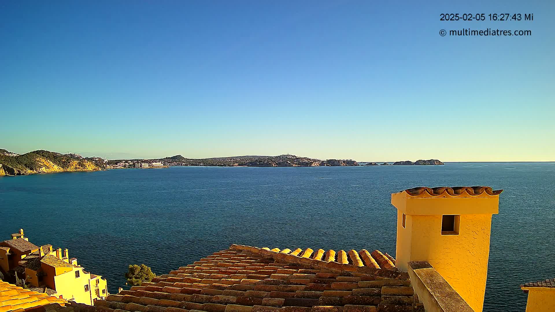 A coastal view on a clear, sunny day shows a terracotta roof, a yellow chimney, and a calm sea extending to a distant shoreline.