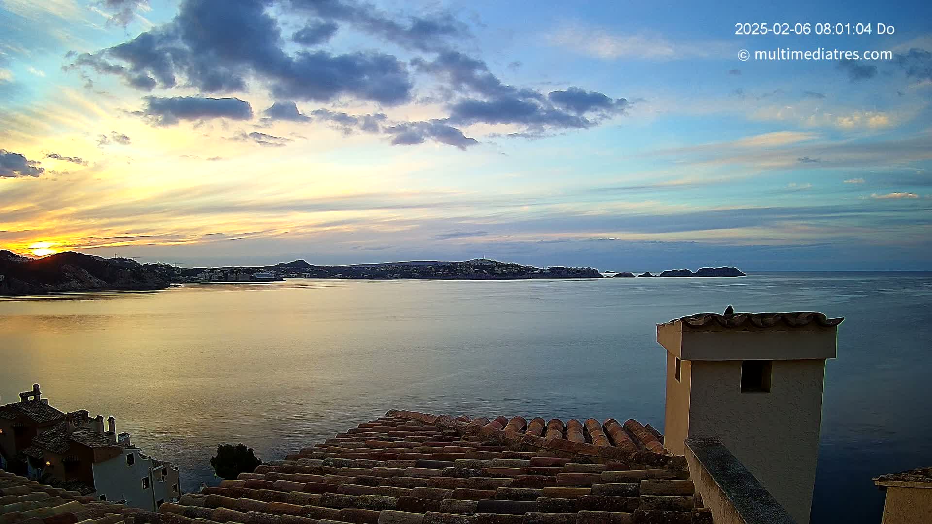 A coastal sunrise scene with a partly cloudy sky is viewed from a rooftop with terracotta tiles, overlooking a calm body of water and distant hills.
