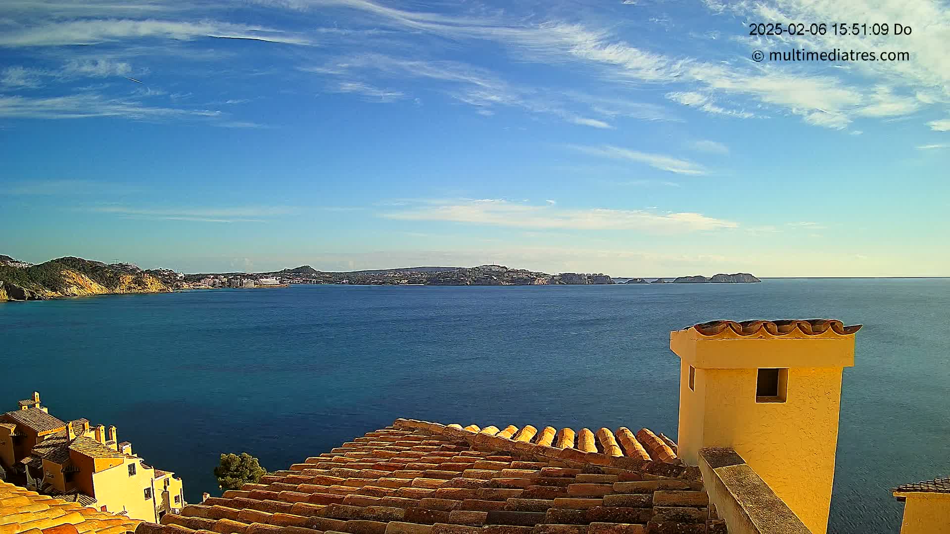 A view from a terracotta roof shows a calm, blue sea under a mostly sunny sky with a few wispy clouds, and distant hills and coastline.