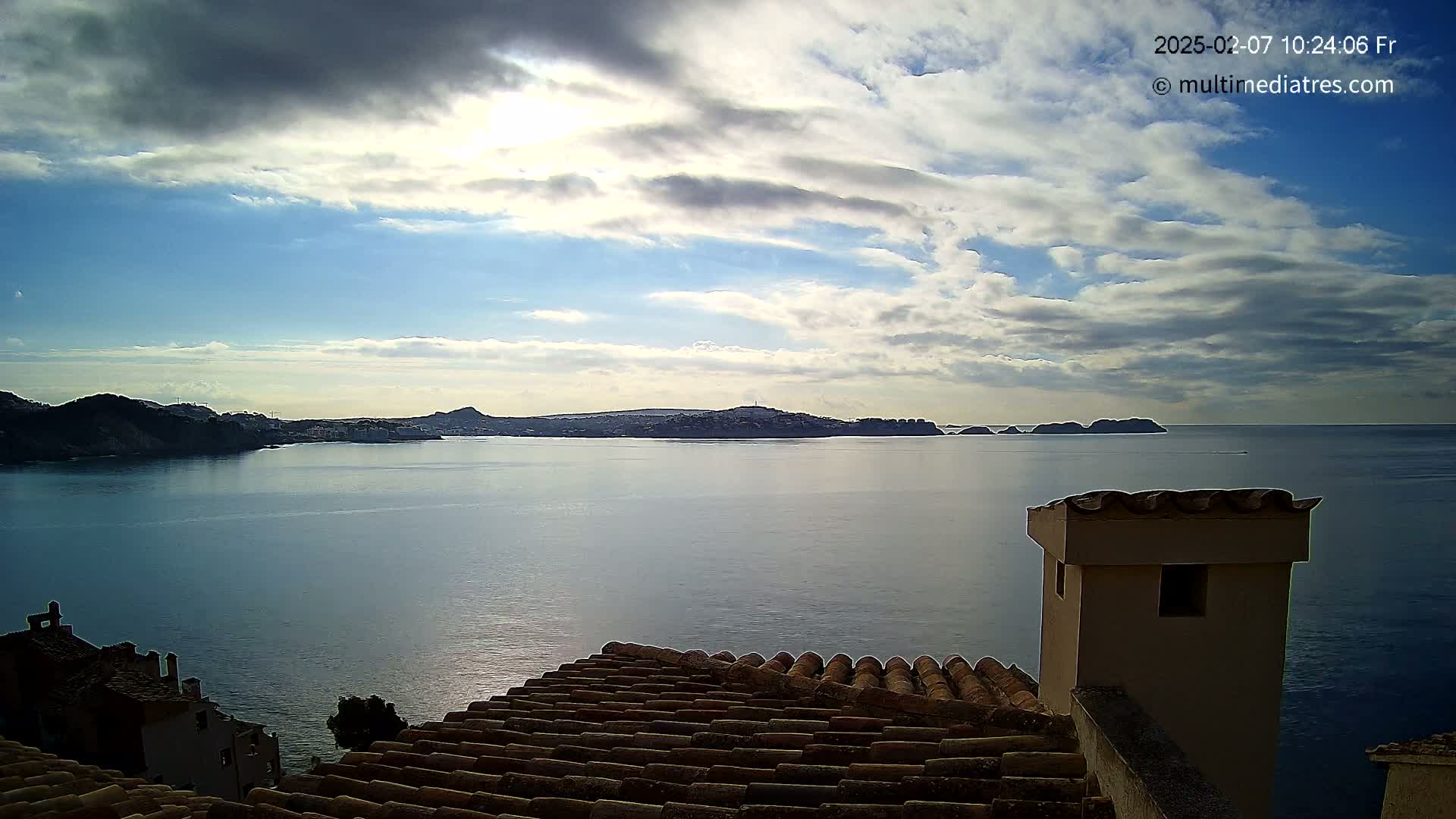 A partly cloudy sky overlooks a calm bay with distant hills and a tiled roof in the foreground.